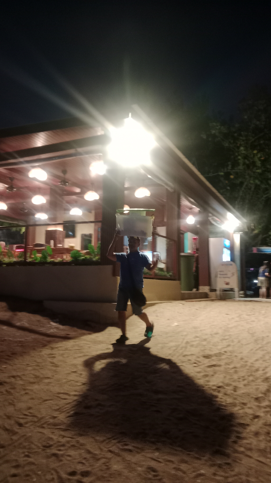 A man is walking across a sandy area in front of a building at night. He is wearing a blue t-shirt and dark shorts, and he is carrying a large rectangular object on his head. The building appears to be a restaurant or bar, with multiple lights illuminating the interior and exterior. The man is casting a long shadow on the sand. In the background, other people and buildings are visible, suggesting a populated area. The scene is likely Kuta, Indonesia, a popular tourist destination known for its beaches and nightlife.