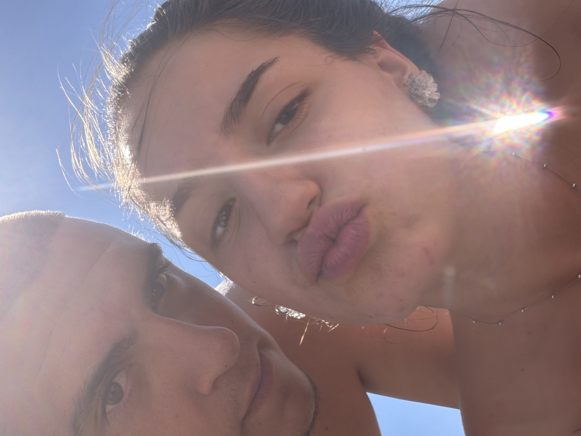A close-up, low-angle outdoor selfie features a couple against a bright blue sky, suggesting a sunny day. The sun's glare creates lens flares and bright highlights. The man's face is partially visible on the left, with his eyes looking towards the camera. The woman is positioned in the center and right of the frame, with her lips pursed and her eyes looking towards the camera. She is wearing a floral-shaped earring in her right ear and a delicate silver necklace. Her hair is windswept. The overall lighting indicates it is daytime, and the presence of the sky suggests an open environment. The scene's content and lighting are consistent with a beach or outdoor recreational area, which aligns with the known characteristics of Primorsko, Bulgaria, a coastal town known for its sunny beaches.
