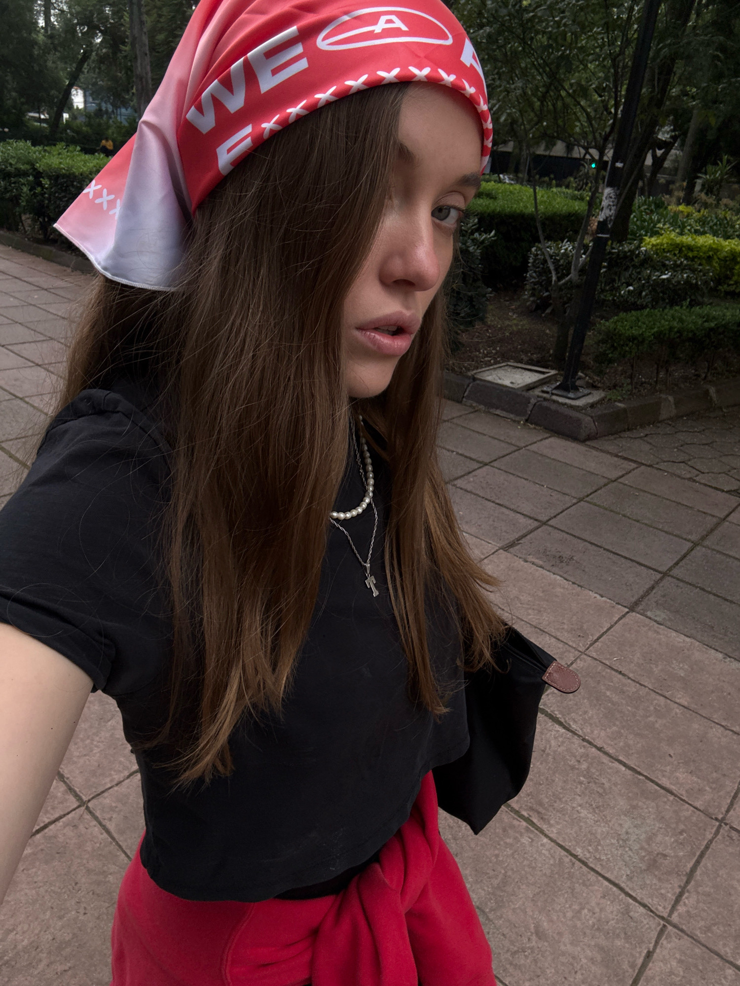 This is a selfie of a young woman in Mexico City, Mexico. She has long, brown hair and is wearing a red bandana with white text, a black t-shirt, and a red sweatshirt tied around her waist. She is also wearing a pearl necklace with a silver pendant. The woman is standing on a paved walkway in a park-like setting with trees and manicured bushes. The ground is paved with square tiles, and there are some streetlights in the background. The lighting suggests it is daytime, possibly late afternoon or early evening. The woman has a serious expression and is looking directly at the camera. The bandana has the word "WE" clearly visible, along with some other partially obscured text and patterns. The overall impression is a casual, urban outdoor scene.