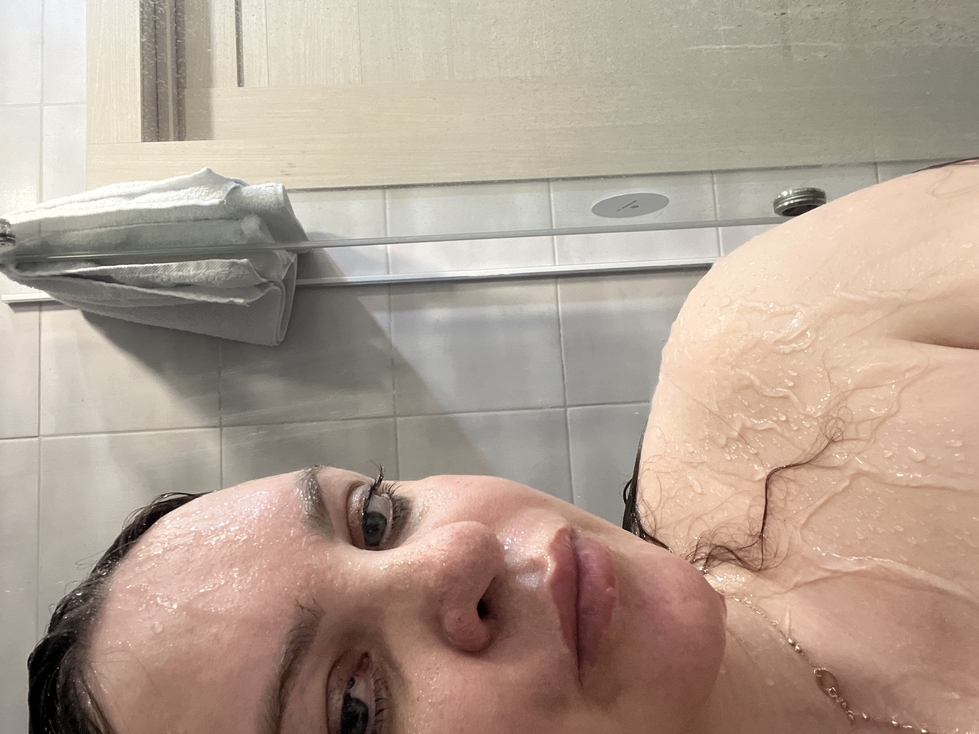 The image is a close-up selfie of a woman with wet skin, likely taken in a bathroom or shower. Her face fills the lower left quadrant of the frame. Beads of water cover her forehead and face. She is fair-skinned with dark hair. The upper right quadrant shows part of her wet shoulder and neck, also covered in droplets. A gold necklace is partially visible around her neck. In the background, a white towel is draped over a rack against a tiled wall. A bathroom fixture, possibly a light or vent, is visible on the upper wall. There is no visible location information to suggest it was taken in the city of Noviy, Russia.