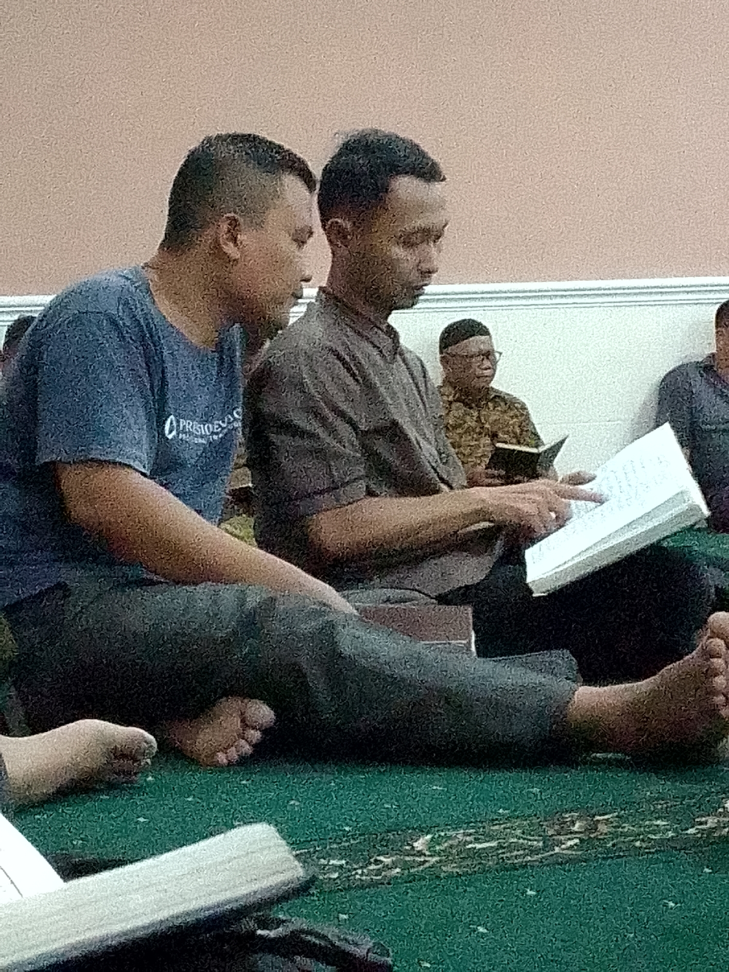 Two Indonesian men are sitting on a green carpeted floor, likely within a mosque or community center in Kertajaya, Indonesia. The man on the left, wearing a blue t-shirt with an emblem and dark pants, is looking towards the right. The man on the right, wearing a brown collared shirt and dark pants, is looking down at an open book held in his hands. His right hand is on the book, with his index finger pointing to a page. Another man, wearing glasses and a patterned shirt, is seated further back, holding a smaller open book. The background features a light-colored wall. The overall setting appears to be an indoor communal gathering.