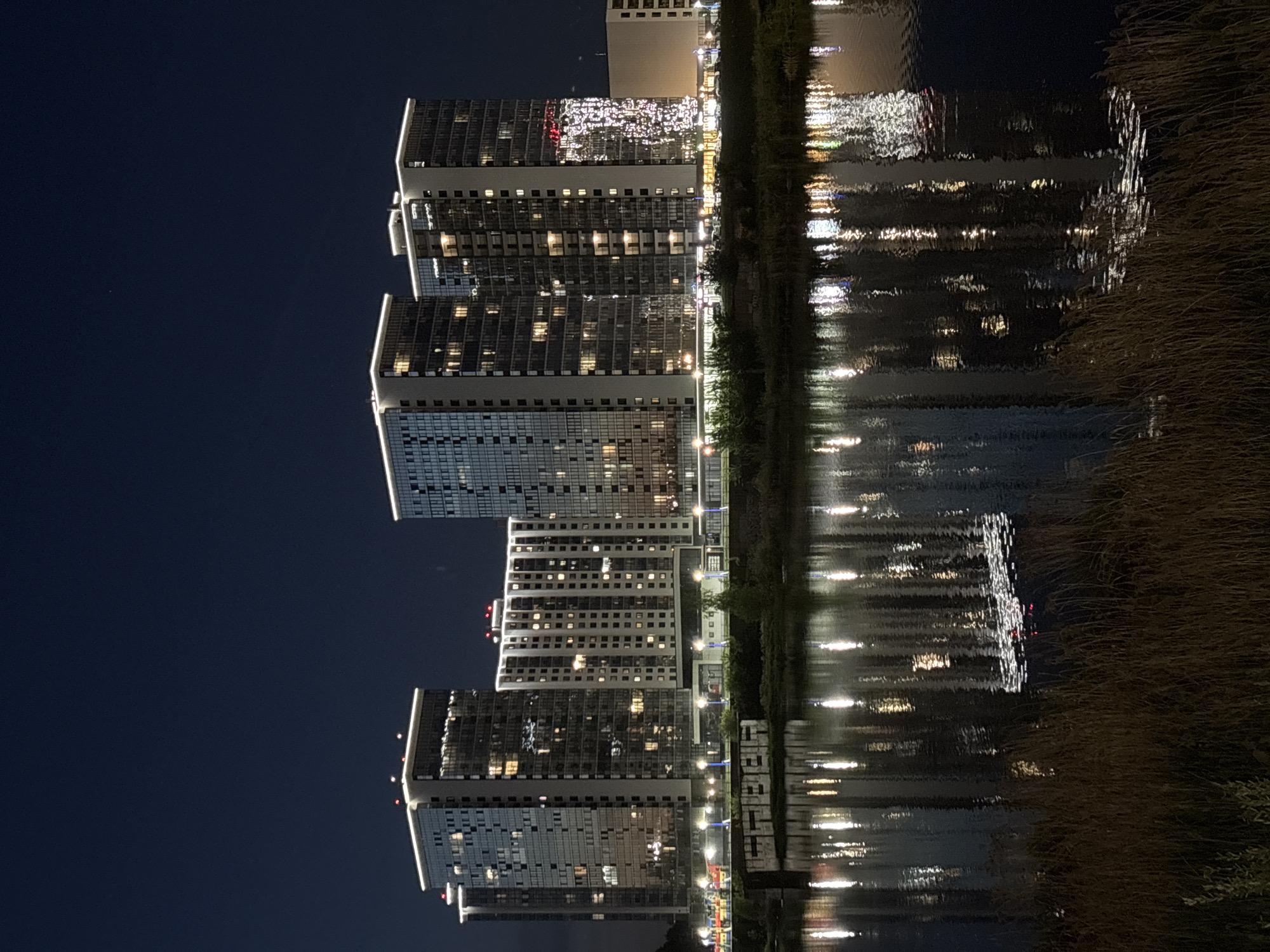 The image shows a nighttime view of modern high-rise apartment buildings reflected in a body of water, likely a lake or river. The buildings have a mix of dark and lit windows, suggesting residential occupancy. Along the water's edge, there's vegetation. Given the prompt, this scene is likely located in Naberezhnye Chelny, Russia.