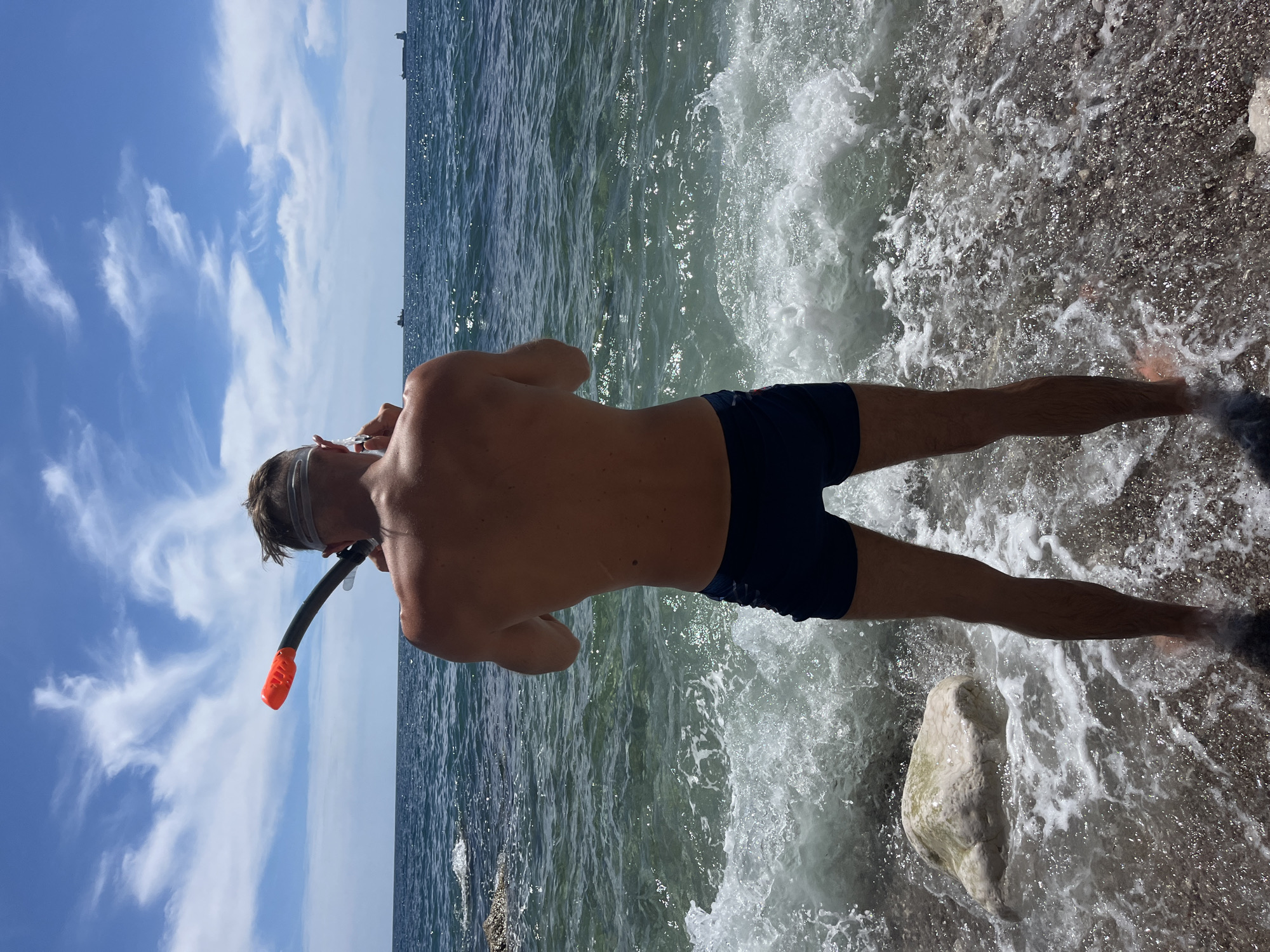 The image depicts a person, viewed from behind, standing at the edge of the sea in Bar, Montenegro. The individual is wearing dark swimming trunks and a head strap with a snorkel that has an orange mouthpiece. They are facing the water, which is clear and shows small waves breaking on a pebble beach. In the distance, two cargo ships are visible on the horizon under a partly cloudy blue sky. The sunlight creates reflections on the water's surface.