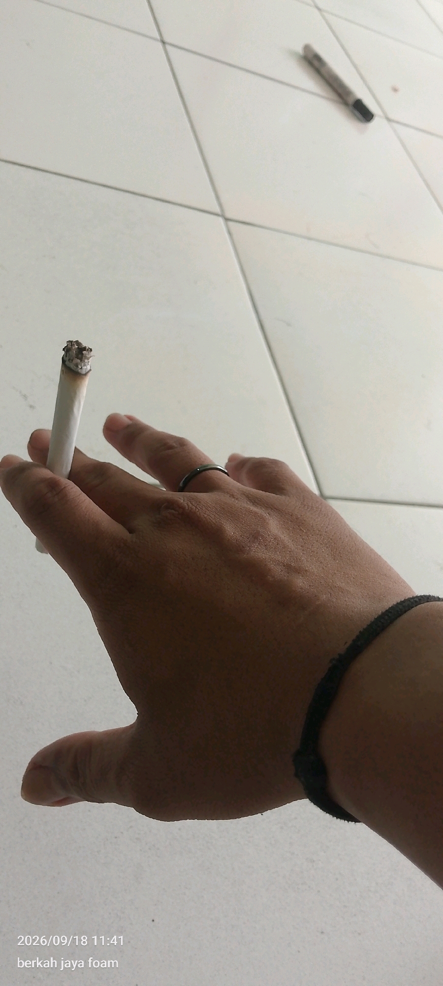 The image displays a close-up view of a hand holding a lit cigarette, with a dark ring on one finger and a black bracelet on the wrist. The hand is positioned over a tiled floor, which appears to be white and laid out in a grid pattern. In the background, on the floor, is a writing instrument, possibly a pen. The timestamp "2026/09/18 11:41" and text "berkah jaya foam" are superimposed in the lower-left corner, indicating a date, time, and potentially a business name or location. The scene is lit from above, casting soft shadows. The environment is indoors, as suggested by the tiled floor and the presence of a pen. There are no discernible landmarks or contextual clues that would specifically place this scene in Kincang Wetan, Indonesia.