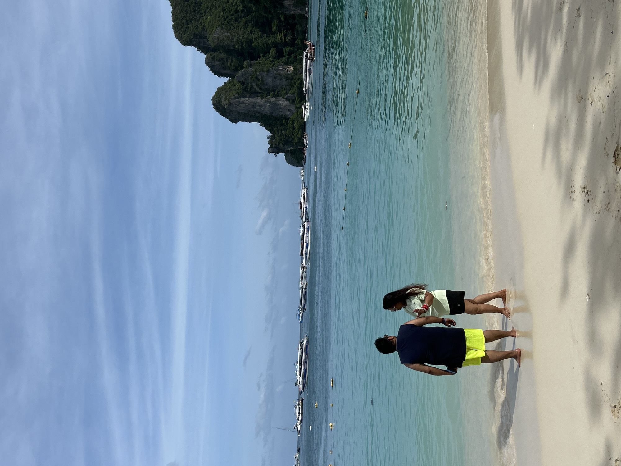 A man and a woman stand facing the ocean on a sandy beach in Ao Nang, Thailand. The man, on the left, wears a dark blue sleeveless shirt and bright yellow shorts. The woman, on the right, is dressed in a light green t-shirt and black shorts. They are positioned near the water's edge, with gentle waves lapping at their feet.
The ocean is a clear turquoise color and dotted with numerous boats, including speedboats and what appear to be longtail boats. In the background, towering limestone karsts covered in lush green vegetation rise dramatically from the water, characteristic of the Phi Phi Islands area, which is accessible from Ao Nang. The sky is a pale blue with scattered white clouds. The overall scene depicts a serene tropical beach setting.