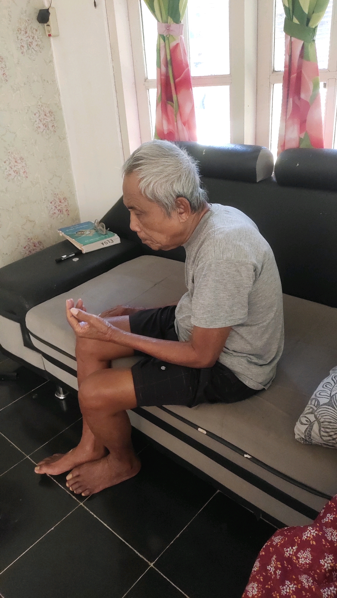 An elderly Indonesian man with gray hair sits on a couch in what appears to be a residential setting. He is wearing a gray t-shirt and black shorts. His left leg is crossed over his right, and his hands are clasped in front of him. The couch is dark gray and beige, and it is placed in front of a window with pink and green floral curtains. A book and a pen are on the couch to the left of the man. The floor is tiled in black and white. The overall lighting suggests it is daytime.