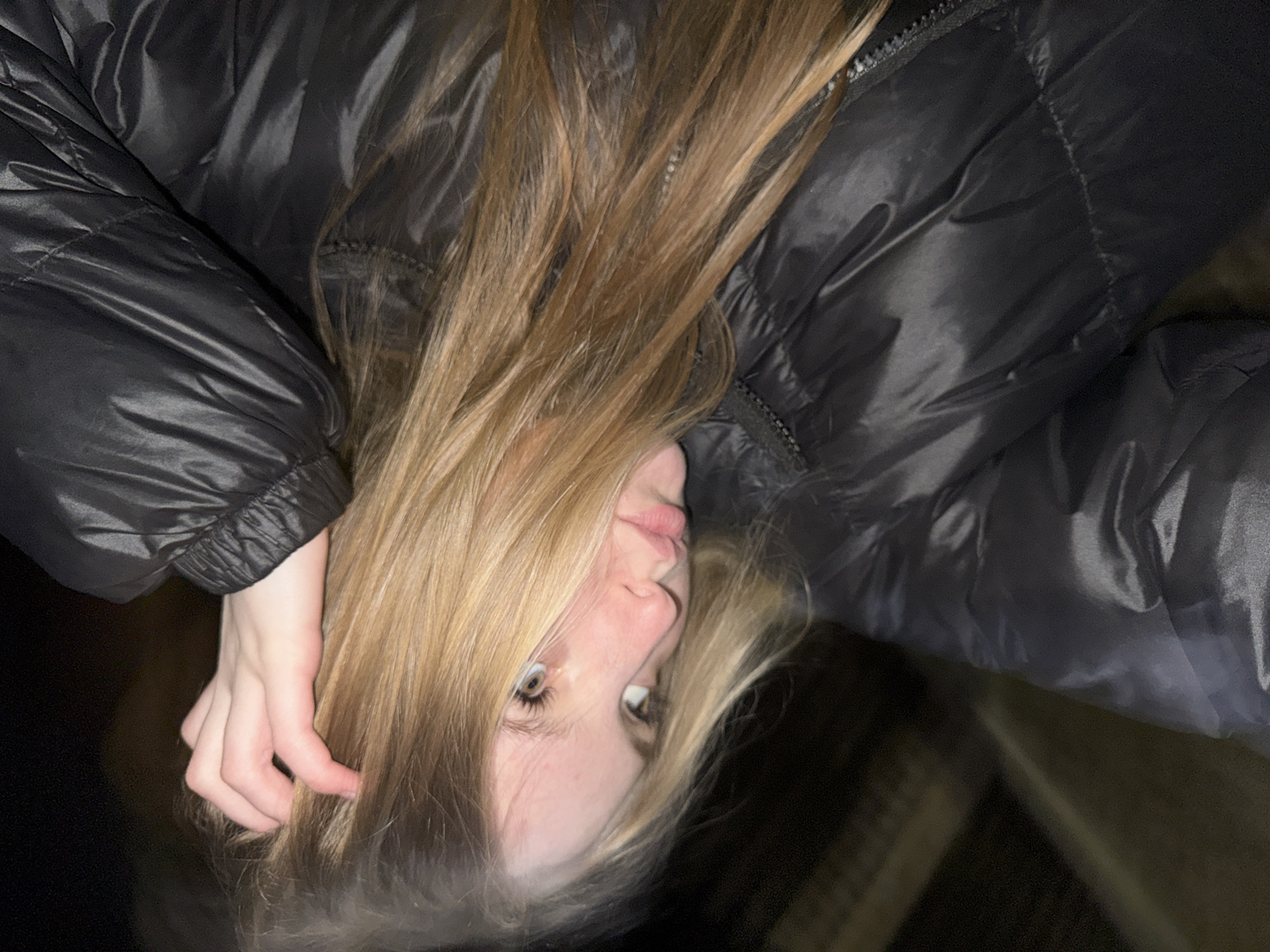 The image presents a woman with long, blonde hair wearing a black puffy jacket. The orientation is such that she appears upside down in the frame. One hand is visible, placed near her hair. The background is dark, obscuring details of the location. The scene lacks any specific details linking it to Zhigalki, Russia.