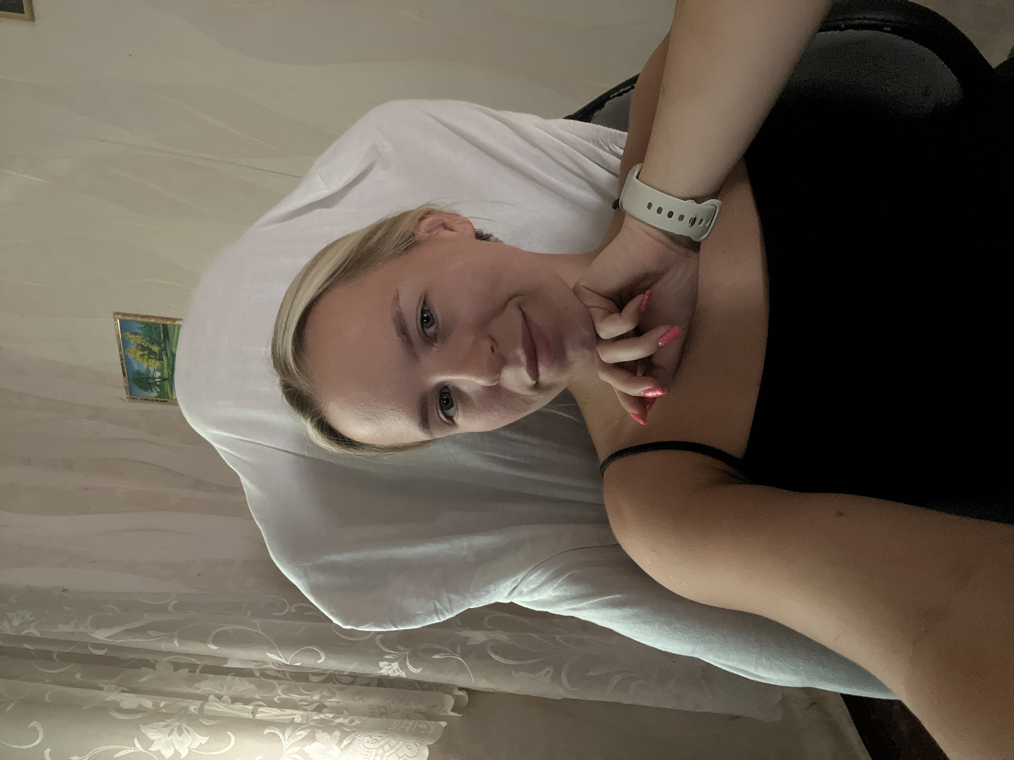 A young woman with blonde hair is sitting in a chair, her chin resting on her hand, looking at the camera. She is wearing a black tank top and a white smartwatch on her left wrist. The chair is draped with a white cloth. In the background, there are sheer curtains and a small framed picture on the wall. The overall setting appears to be an indoor, domestic environment.
