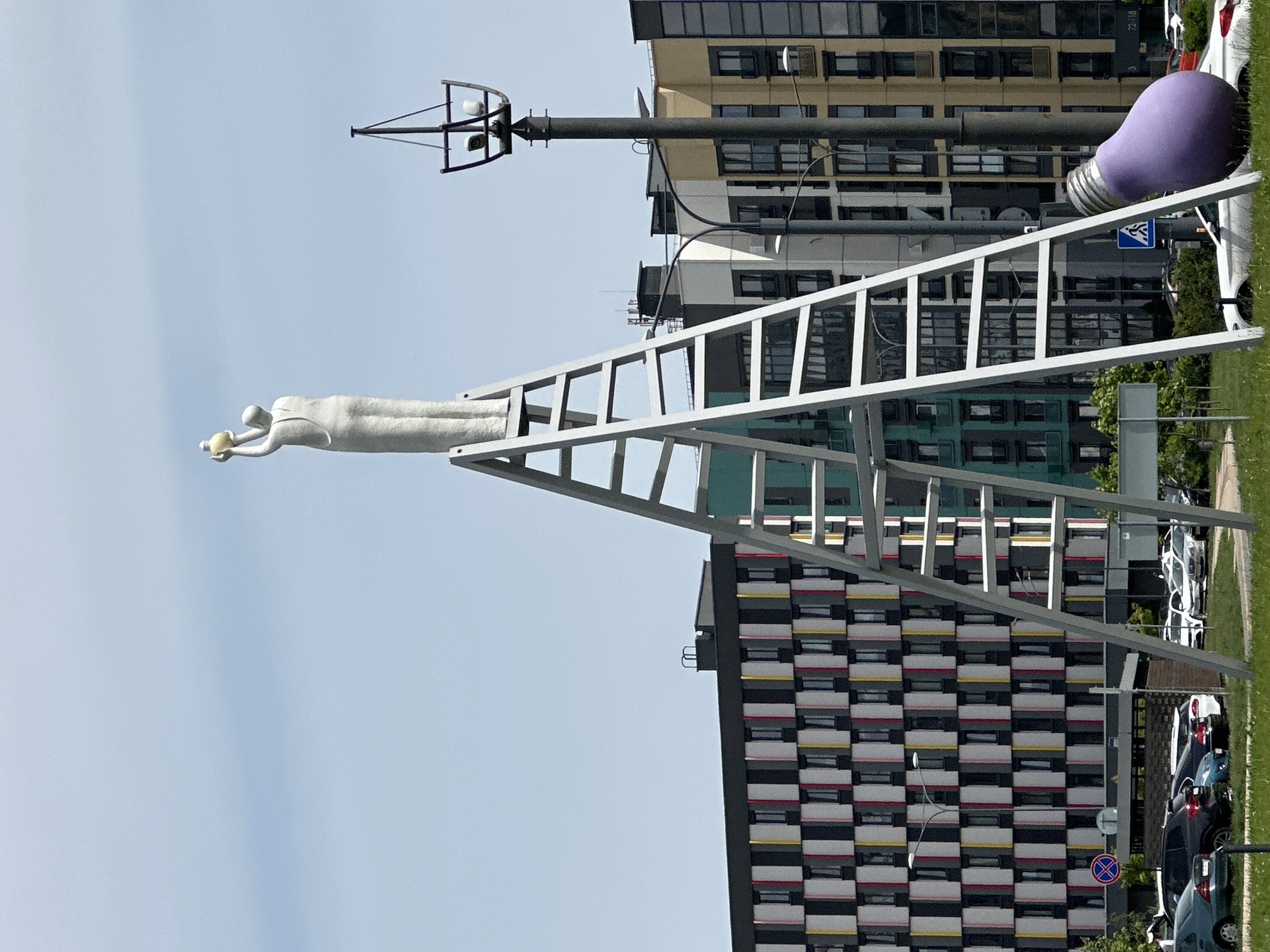 The image depicts a public artwork in the city of Бараўлянскі сельскі Савет, Belarus. The artwork consists of a white ladder that is supported by a metal frame. At the top of the ladder, a white statue of a human figure is positioned horizontally, holding an object. At the opposite end of the ladder, there is a large purple lightbulb. In the background, there are several apartment buildings with a checkered pattern. A light pole extends upwards from the base of the frame. There is a line of cars parked adjacent to the artwork. Grass is present in the background. The sky is light blue.