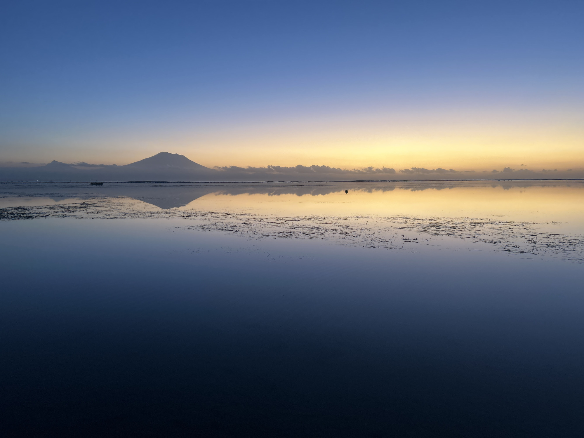 The image captures a serene dawn over a body of water in Denpasar, Indonesia, likely near the coast or a large river mouth, given the presence of a distant, silhouetted volcano. The sky transitions from a deep blue at the top to a soft gradient of orange and yellow near the horizon, indicating the early stages of sunrise. A prominent volcano, Mount Agung, is visible in the far distance, its peak partially obscured by a thin layer of clouds. The calm water acts as a mirror, reflecting the sky and the volcano with remarkable clarity, creating a symmetrical and tranquil scene. The foreground of the water is dark and deep, while closer to the horizon, a band of lighter, shallower water is visible, possibly indicating mudflats or areas with aquatic vegetation. A small, dark object, possibly a buoy or a small boat, is present in the middle ground of the water. The overall impression is one of peace and natural beauty, characteristic of sunrise over coastal areas in Bali.
