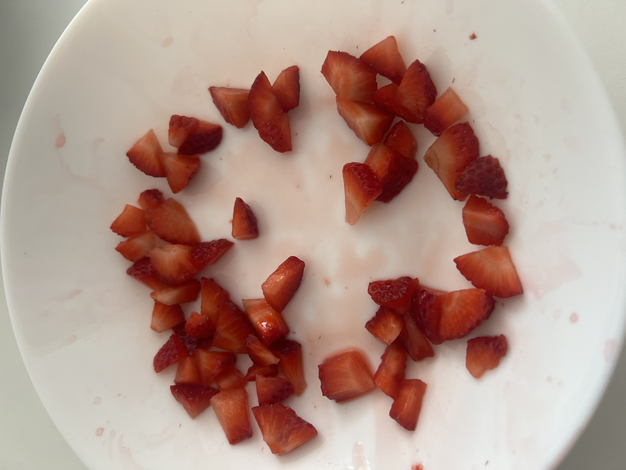 A white, round plate contains irregularly arranged, diced strawberries. The plate shows residue of strawberry juice. The location and date are not discernible from the image. No people are visible.