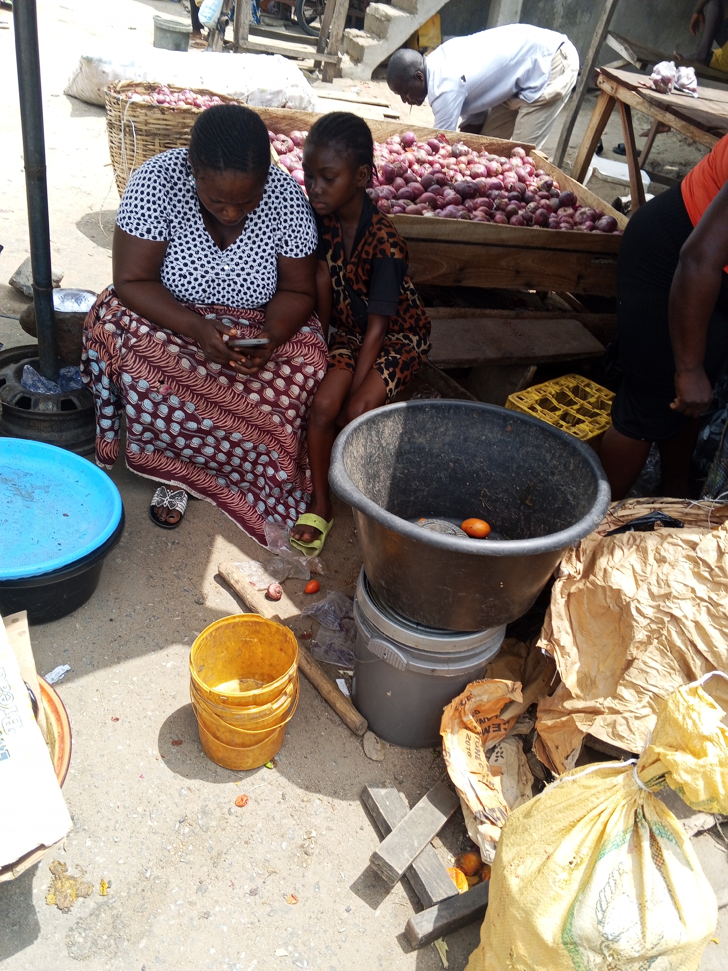 In Zango-ndaji, Nigeria, an outdoor market scene unfolds with a seated woman, adorned in a white and black polka-dot top and a patterned skirt, gazing at a smartphone. Beside her, a young girl in a brown animal-print shirt sits attentively. A man in a white shirt is seen behind them sorting through a large assortment of onions on a wooden stand. To the foreground, a black plastic tub with an orange fruit rests on stacked grey buckets. A stack of yellow plastic buckets is nearby, and a yellow sack lies to the right, partially obscured by brown paper. The ground shows signs of market debris.