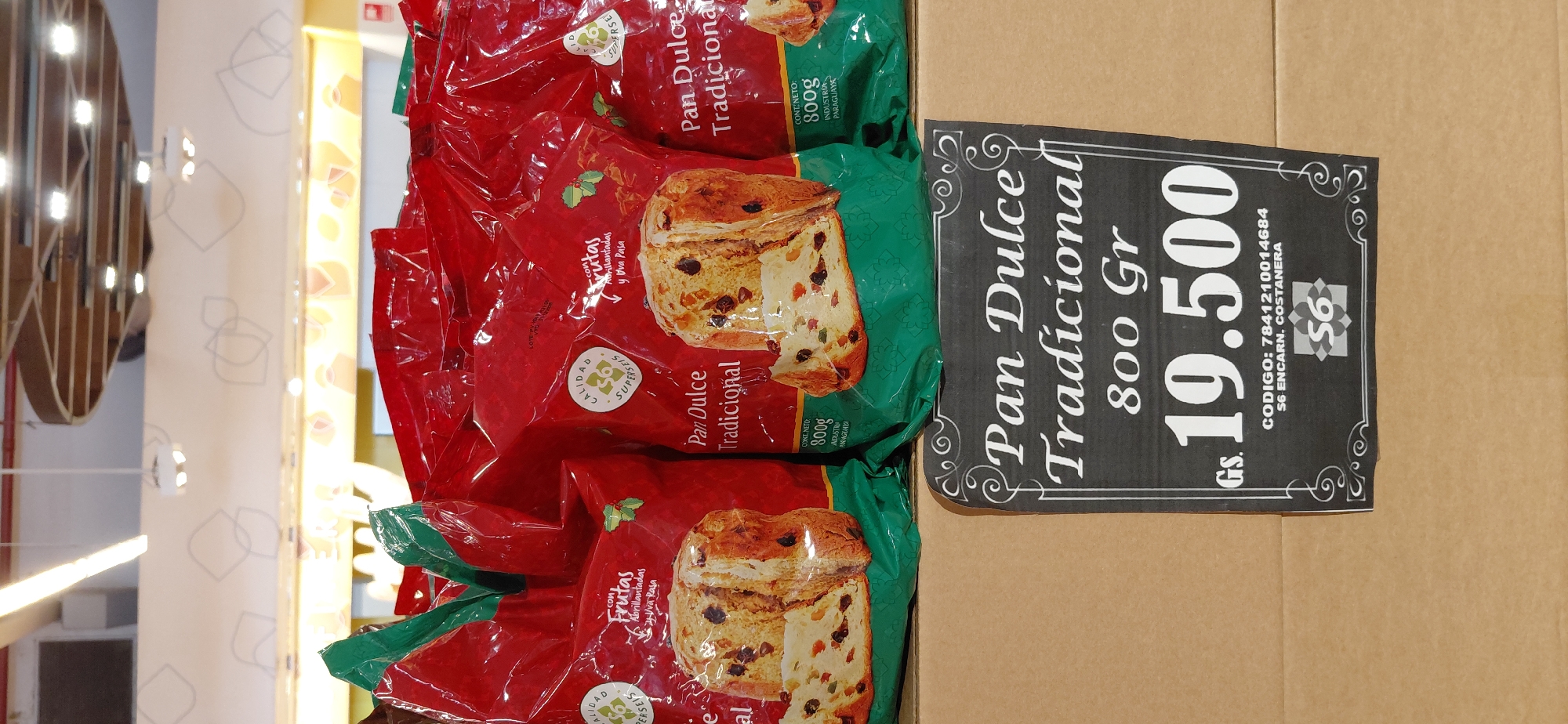 A close-up shot shows a display of Pan Dulce Tradicional, a traditional sweet bread, in a supermarket. The bread is packaged in bright red bags with green accents, featuring images of sliced bread with visible fruit pieces like raisins and candied peel. Several bags are stacked, with the foreground showing two prominent bags. Below the bread display, a black price tag is prominently displayed, indicating "Pan Dulce Tradicional 800 Gr" and the price "19.500" Gs. (Guaraníes), the currency of Paraguay. The price tag also includes a code "7841210014684" and "S6 ENCARN. COSTANERA," identifying the location as Encarnación, Costanera. The background suggests a retail environment with shelving and lighting typical of a supermarket.