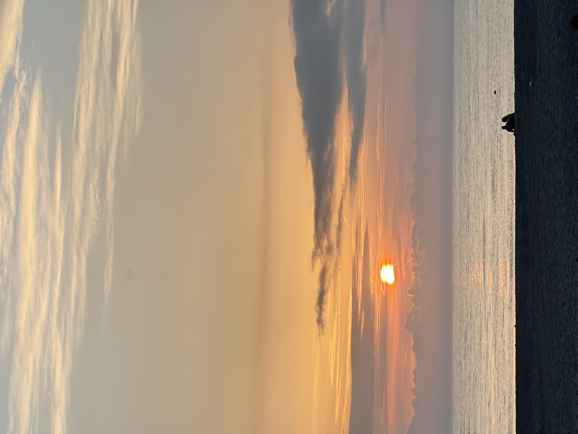 The image captures a sunset over the sea in Sochi, Russia. The sun, a bright orange orb, is partially obscured by clouds near the horizon, creating a dramatic light effect. The sky transitions from a light blue with wispy clouds on the left to a warm, golden hue towards the center where the sun is setting.
To the right, the sea reflects the sunset's glow, with a couple of people standing close to the edge of a dark structure or wall that borders the water. The water is calm, with subtle ripples reflecting the light.