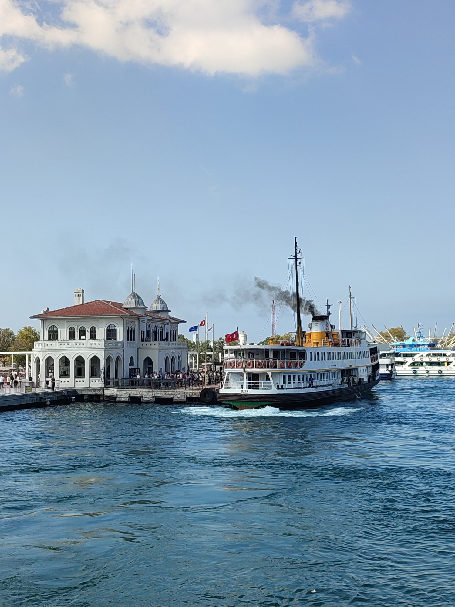 In Kadıköy, Turkey, a ferry departs from a pier next to a white building with a red roof. The building has arched windows and two domes. The ferry, emitting black smoke from its smokestack, is white with orange life preservers and a Turkish flag. Several other boats are visible in the background. The water is a deep blue, and the sky is clear with scattered white clouds. A crowd of people is gathered on the pier.