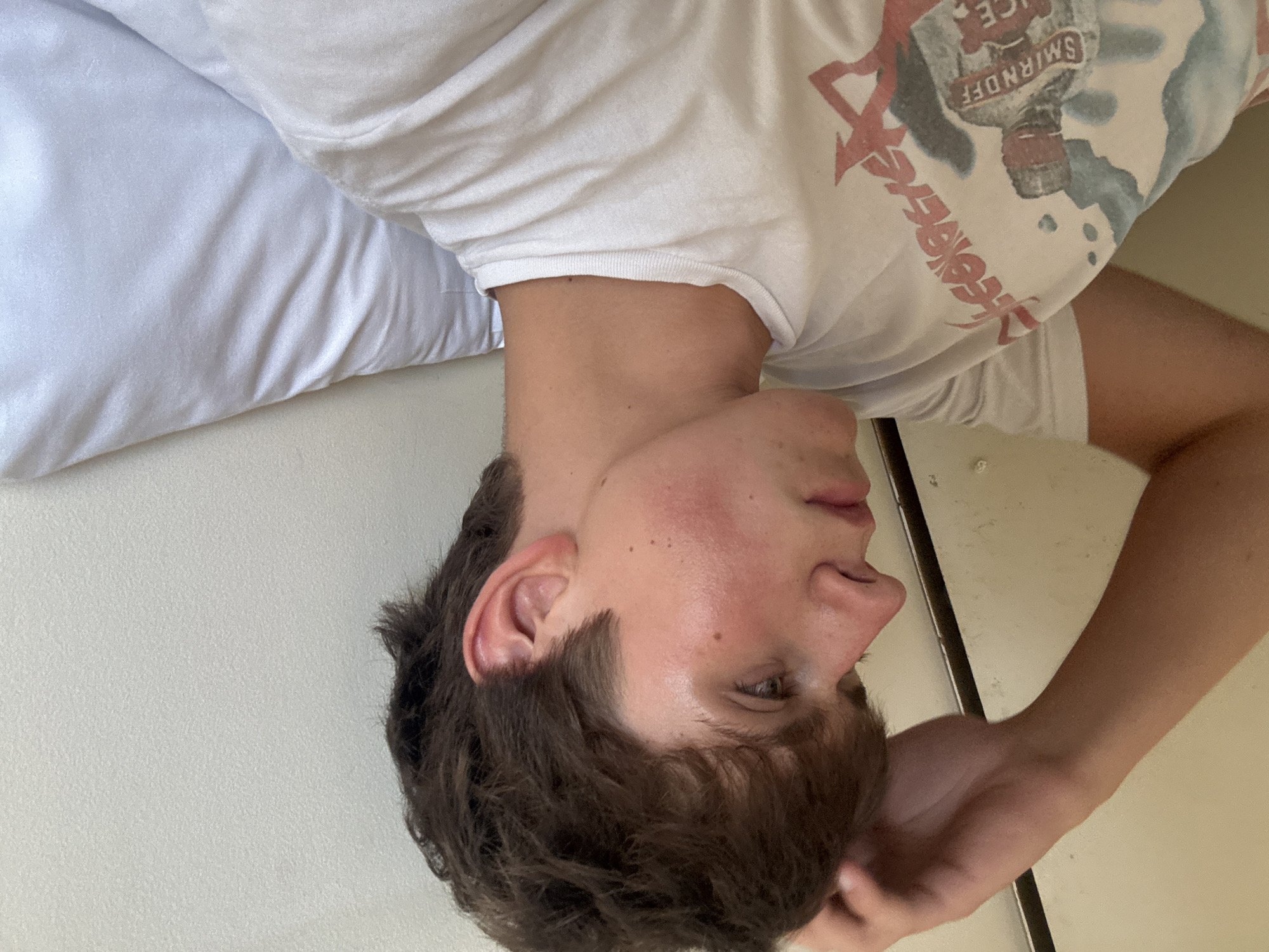 An unidentified individual with short brown hair is lying in bed, propped up by pillows. The person is wearing a white t-shirt with a graphic print. The individual's right arm is raised, with their hand resting on their head. The image is taken indoors, and the background consists of a wall and bedding. There is no indication of the location being in London, United Kingdom.
