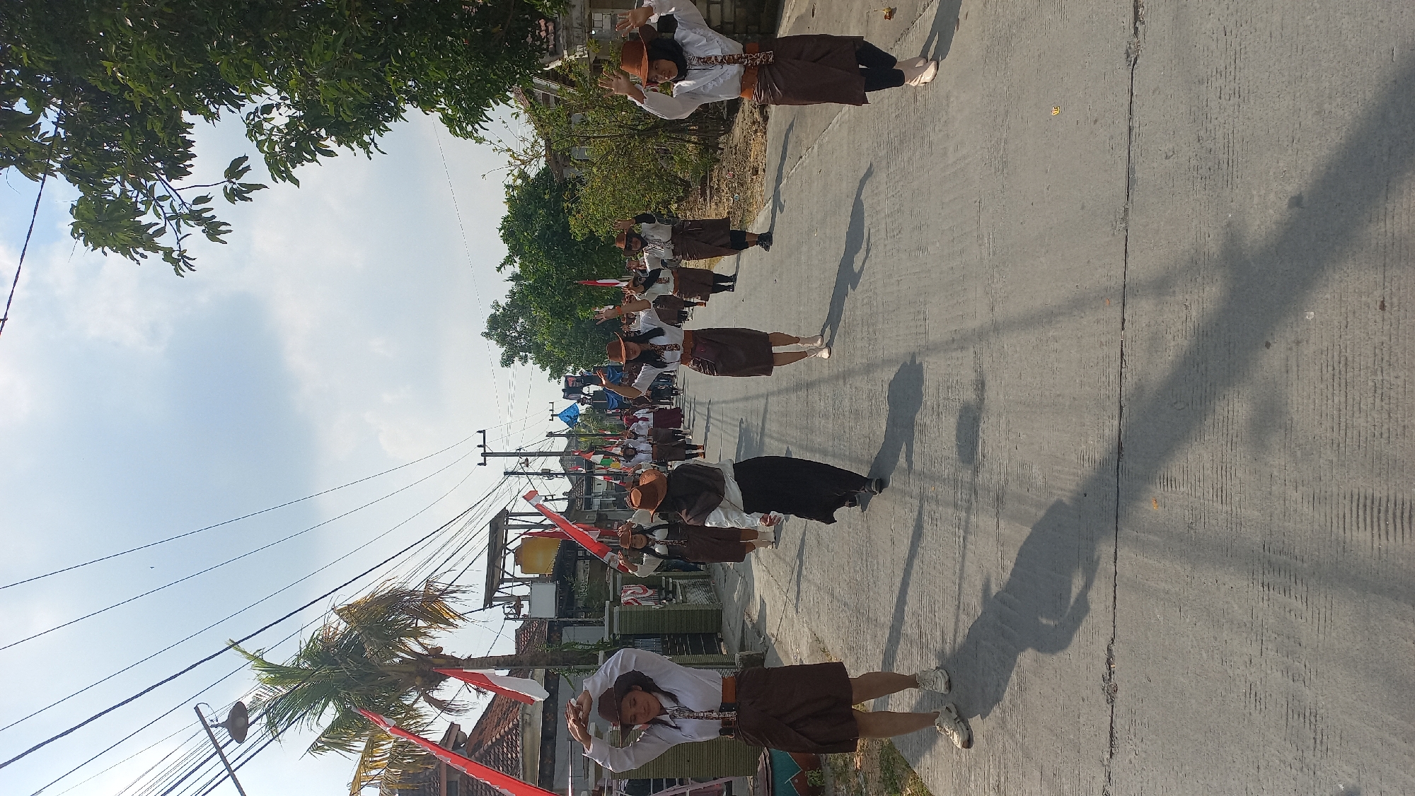 In Kedungmegarih, Indonesia, a group of young women in matching brown skirts and white shirts, accessorized with brown hats and belts, are participating in a parade on a paved road. They are performing a synchronized dance with their arms raised, their movements suggesting a spirited performance. The road is lined with houses and trees, with Indonesian flags displayed prominently on poles and houses, indicating a national celebration. The sky is partly cloudy, with sunlight casting long shadows on the road, suggesting it is late afternoon.