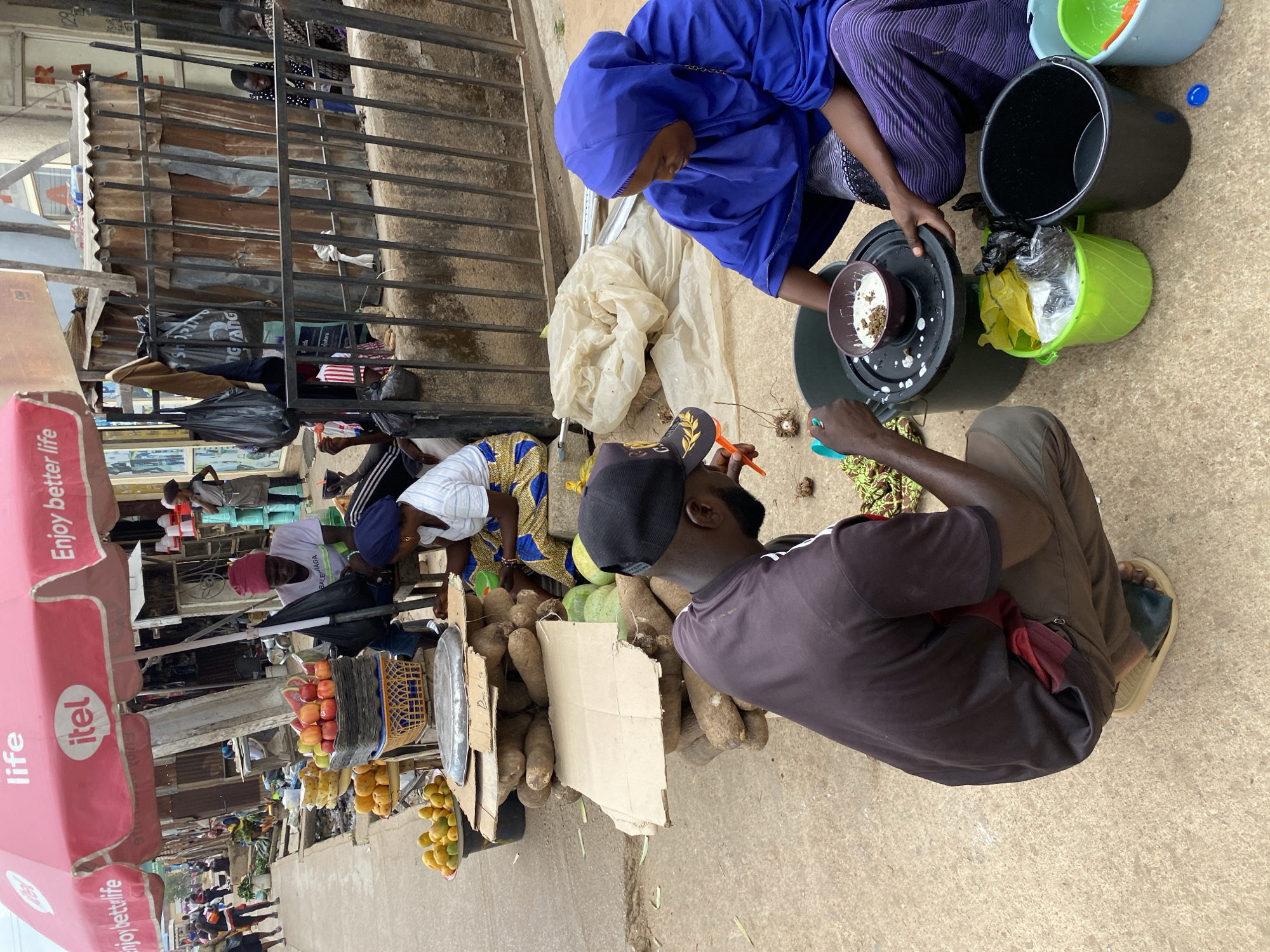 Here's a detailed description of the image:
**General Overview:**
The image captures a street scene in what appears to be Bukuru, Nigeria, with a focus on two individuals engaged in food preparation or sale. The environment includes elements of a market or roadside commerce.
**People:**
* **Two individuals:** A woman wearing a blue hijab and a man with a black cap are the central figures. They are seated on the ground, working together.
* **Other people:** Other vendors and pedestrians are visible in the background, suggesting a bustling market setting.
**Subjects/Objects:**
* **Produce:** The primary objects of interest are yams or similar root vegetables, which are piled up. There are also fruits such as mangoes and apples visible.
* **Utensils and containers:** A variety of bowls and buckets are present, suggesting preparation or cleaning activities. One bowl seems to contain food.
* **Market stalls/Shops:** Metallic structures and stalls with produce are observed in the background, indicating a market environment.
* **Advertising:** There's an umbrella bearing the logo "itel life" for mobile phones, indicative of local advertising.
**Actions/Interactions:**
* **Food preparation/Sale:** The man and woman are actively involved in processing food, possibly preparing it for sale. The woman seems to be working with a food item in a bowl. The man appears to be peeling or sorting something using an orange tool.
* **Commerce:** The arrangement of the produce and the presence of containers suggest a commercial activity, likely food vending.
**Environment:**
* **Street/Roadside:** The scene unfolds on a concrete surface, which likely serves as a street or roadside area where informal commerce takes place.
* **Urban setting:** The background reveals buildings and other urban features, further establishing the scene as part of a city.
* **Bukuru, Nigeria:** the style of the stalls and goods point to Nigeria.
