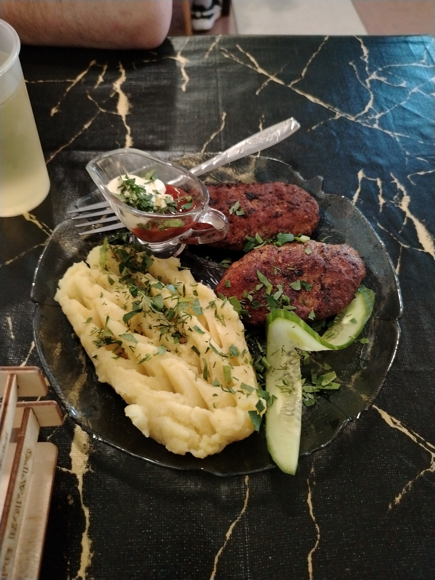 The image shows a plate of food on a table in what appears to be Глебовское, Russia. The plate contains two cutlets, a serving of mashed potatoes, cucumber slices, and a sauce dish with both ketchup and mayonnaise. The mashed potatoes and cutlets are garnished with dill. A fork and spoon rest next to the sauce dish. A glass of liquid, possibly juice or water, is partially visible in the upper left corner. The table has a black marble-like surface with gold veins. A partially visible wooden object is in the lower left. A person is seated at the table.