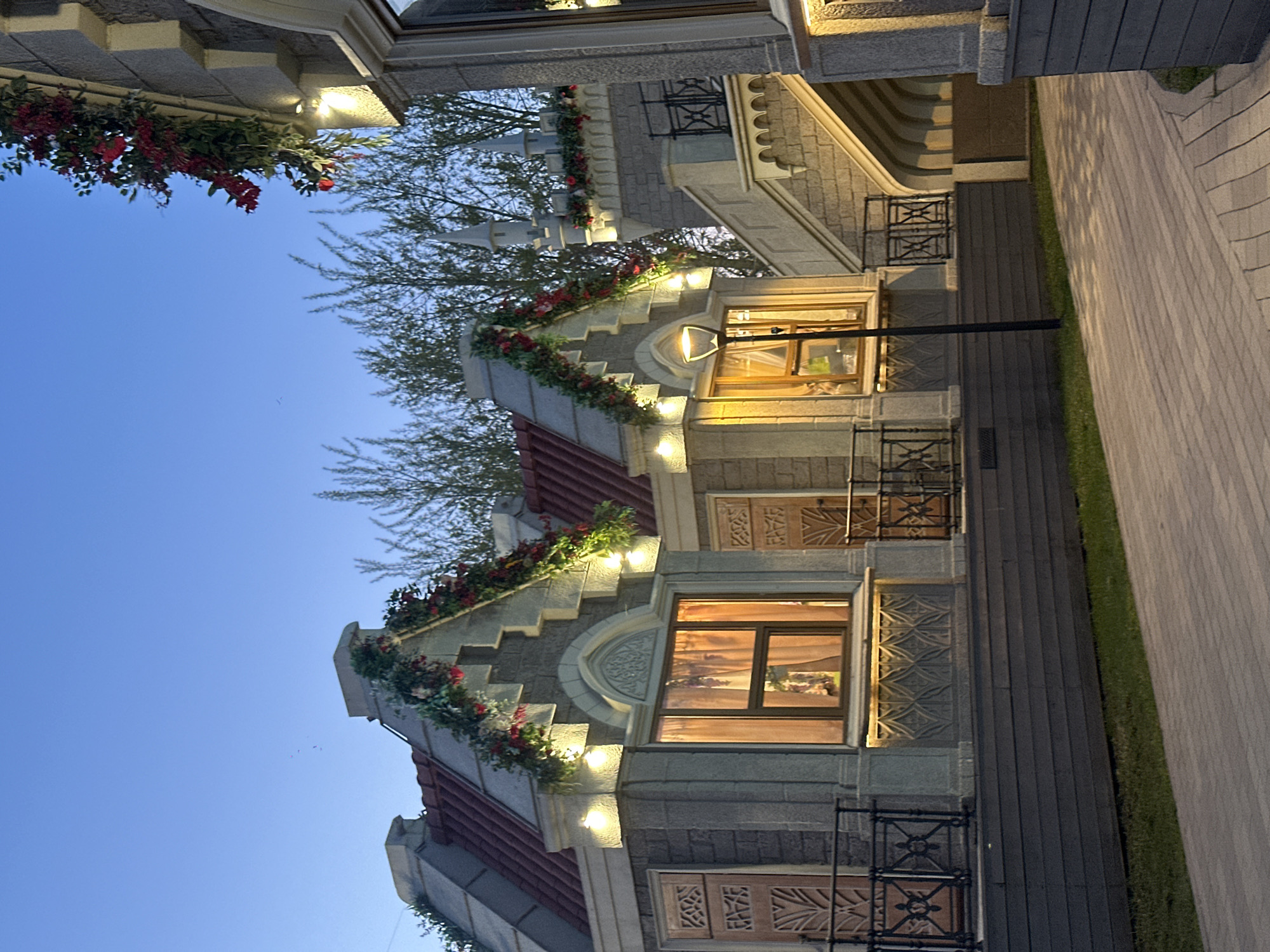 The image shows a building facade, likely in Moscow, Russia, given the architectural style. The building is designed with multiple gables, each adorned with red flowers and greenery. The facade is illuminated by lights placed along the edges of the gables and around the windows. The architecture appears to mimic European styles, possibly Dutch or German, with detailed stone or concrete work. The sky is a clear blue, suggesting it might be dusk or dawn. A paved walkway is visible to the right of the building, separated by a strip of grass. The buildings seem to be part of a larger complex, with other architectural structures visible in the background.