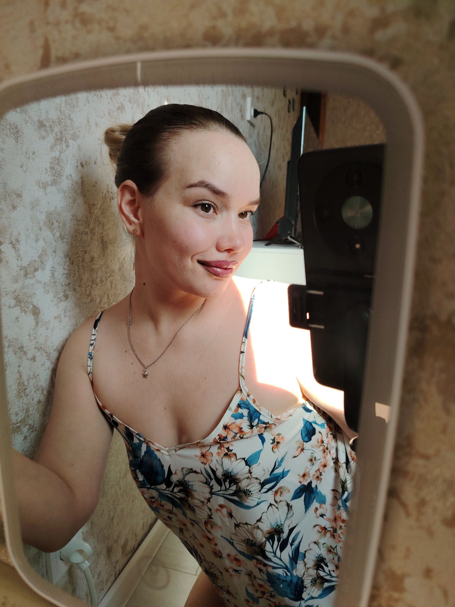 A young adult female is captured in a mirror selfie, located in Марковский, Russia. She is positioned from the chest up, angled slightly to her right, displaying a gentle smile. Her dark hair is pulled back into a bun. She wears a light-colored spaghetti-strap dress adorned with a floral pattern of blue and orange/peach flowers and green leaves. A small gold stud earring is visible in her left ear, along with a silver-toned necklace featuring a star pendant. A prominent light source illuminates her right shoulder and chest. The mirror, through which the image is taken, has a light-colored, rounded rectangular frame. The wall immediately behind the woman is covered in textured, light brown/beige wallpaper. In the background reflected behind the woman's right side, a black electronic device with a silver-colored dial or button rests on a white surface, adjacent to a wall outlet with a black cord plugged in. Another white power outlet with a white cord is visible on the lower left portion of the wall within the reflection. A light-colored floor is visible at the very bottom edge of the frame.