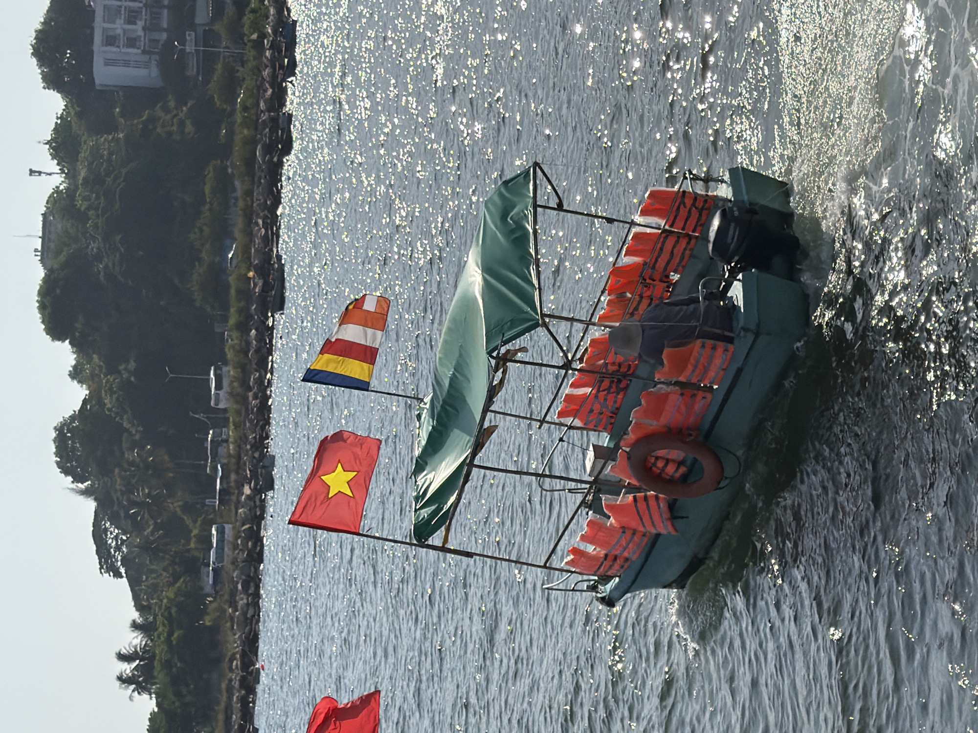 A teal-colored tourist boat with a green canopy is underway on choppy, sun-dappled water. The boat is equipped with a row of orange life vests along its side and a life ring attached to the railing. Two flags are visible at the front of the boat: a red flag with a yellow star, representing Vietnam, and a multi-colored flag with stripes of yellow, red, and blue, likely representing Buddhism. A person wearing a hat and dark clothing is visible on the boat, seated at the helm. The boat has an outboard motor at the stern, which is creating a wake. In the background, a rocky shoreline is lined with palm trees and dense vegetation. Further inland, several buildings, including a multi-story structure, are visible on a hillside. Buses are parked along the shore, suggesting a transportation hub or tourist area. The overall scene depicts a typical day of boat tourism in a coastal Vietnamese city.