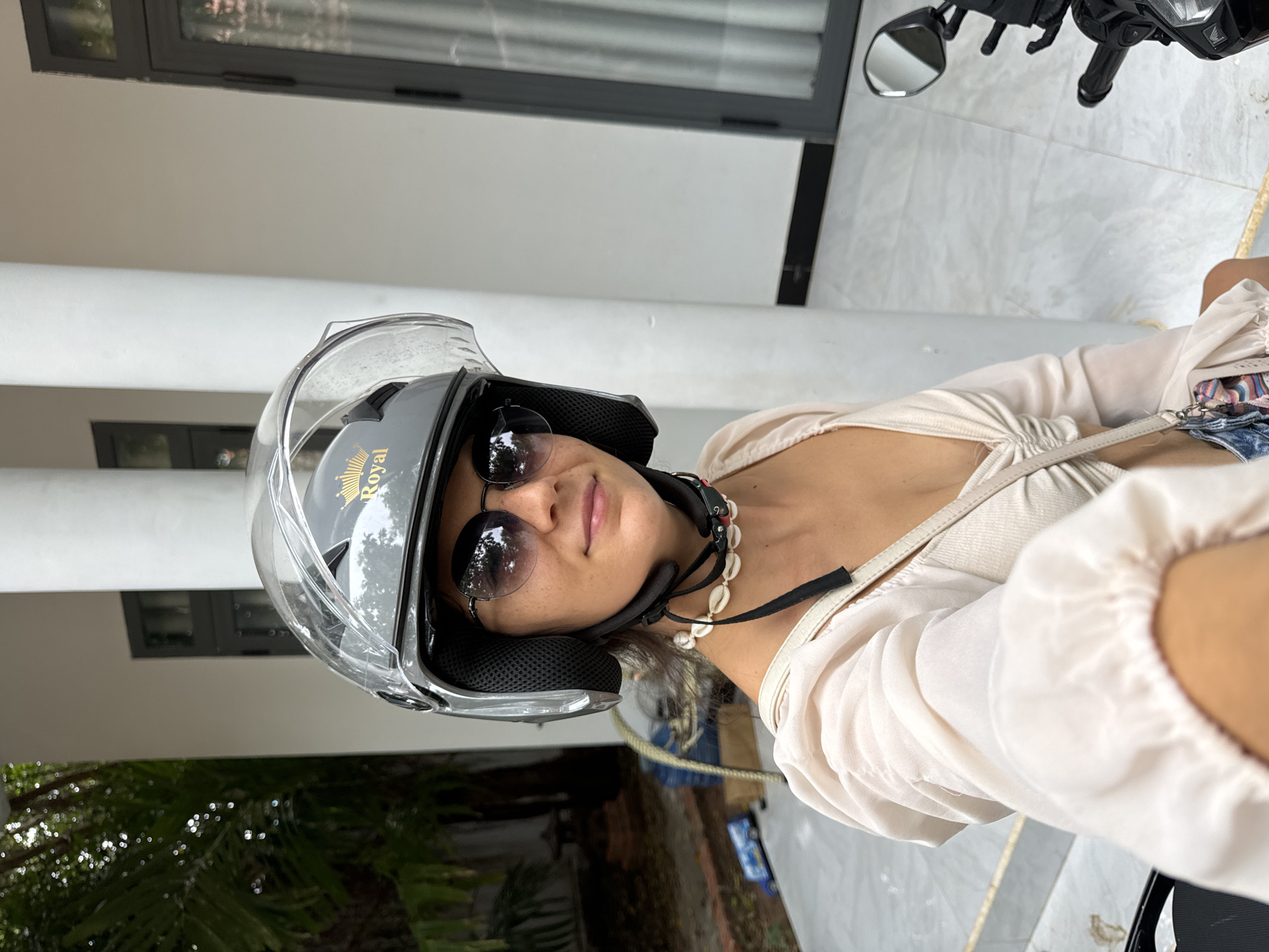 A woman wearing a grey helmet with a clear visor and round sunglasses is taking a selfie. She is wearing a light-colored, off-the-shoulder top with puffed sleeves and a straw bag with a colorful scarf hanging from it. She has a seashell necklace on. The background appears to be a residential area with white pillars, a door, and greenery. The presence of a motorcycle suggests she is about to ride. The scene is consistent with the tropical climate of Mũi Né, Vietnam, known for its beaches and resorts.