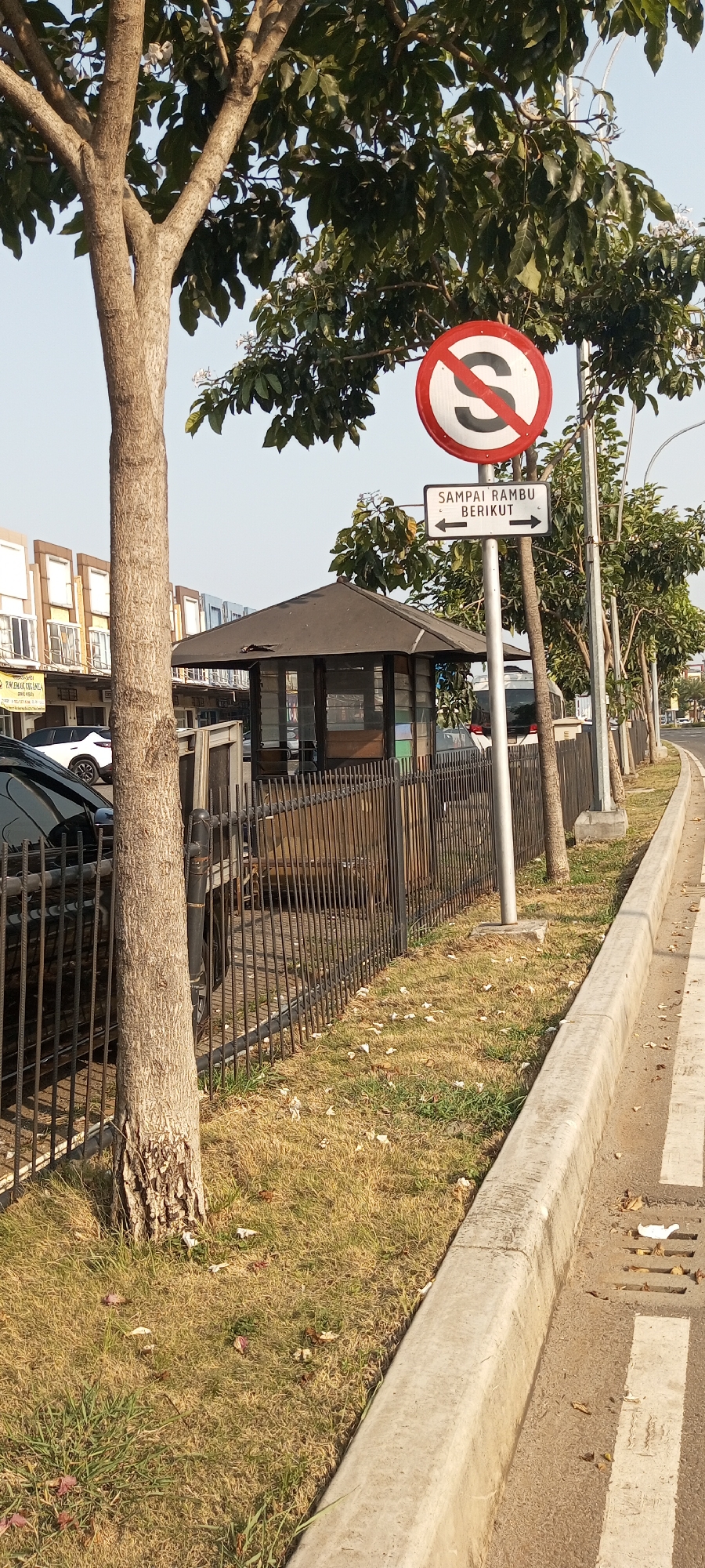 The image captures a street scene in Lambangjaya, Indonesia, featuring a traffic sign, a small guardhouse, a black fence, and a tree. The traffic sign, a red circle with a white background and a black diagonal bar through the letter "S", indicates "no stopping". Below it, a rectangular sign with white text on a black background reads "SAMPAI RAMBU BERIKUT", meaning "until the next sign", with arrows pointing left and right. To the left of the signpost, a small, dark-colored, hexagonal guardhouse with glass windows is visible. A black metal fence runs along the front of the guardhouse and extends to the left, partially obscuring a black car parked behind it. A large tree with a textured, brown trunk and green leaves stands to the left of the guardhouse. The environment appears to be a roadside with a concrete curb and a road marked with white lines on the right side of the image. The ground is covered with dry grass and scattered leaves. The background shows a row of buildings with a light-colored facade and windows. The sky is clear and pale blue. There are no people visible in the image.