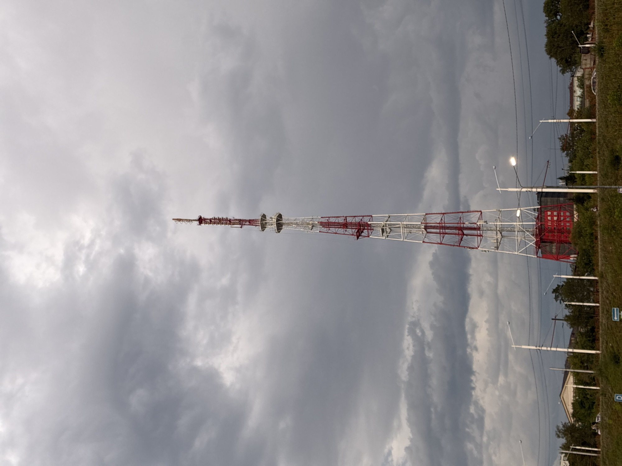 A tall, red and white radio tower dominates the center of the frame, reaching towards a heavily overcast sky. The tower is constructed of a lattice of metal, with sections painted red and white. At its base, a red cylindrical structure is visible. The tower is situated on a slightly elevated grassy area, bordered by a road or pathway in the foreground. Several streetlights are positioned along the path. Behind the tower, a low-lying building with a light-colored roof and trees are visible. The overall scene suggests an outdoor, possibly industrial or semi-rural, environment under a dramatic, cloudy sky, consistent with the weather conditions often observed in Irkutsk, Russia, particularly during transitional seasons. There are no discernible people or animals in the image.