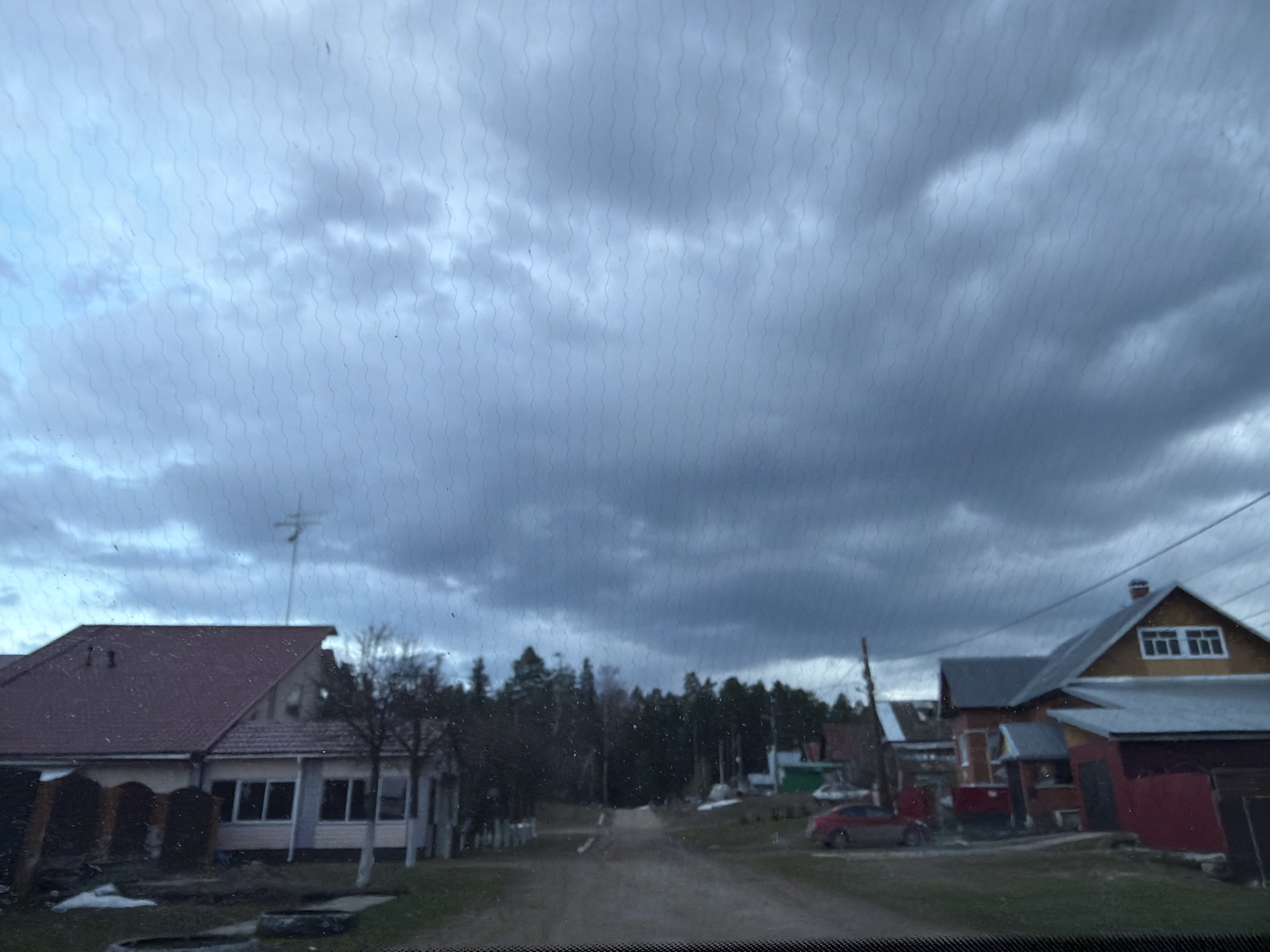 The image shows a view from inside a vehicle, looking out onto a rural street in Chaikovsky, Russia. The sky is heavily overcast with dark, stormy clouds. The road is unpaved. Houses line either side of the road, some with visible roofs. A car is parked on the right side of the street. Trees are visible in the background.