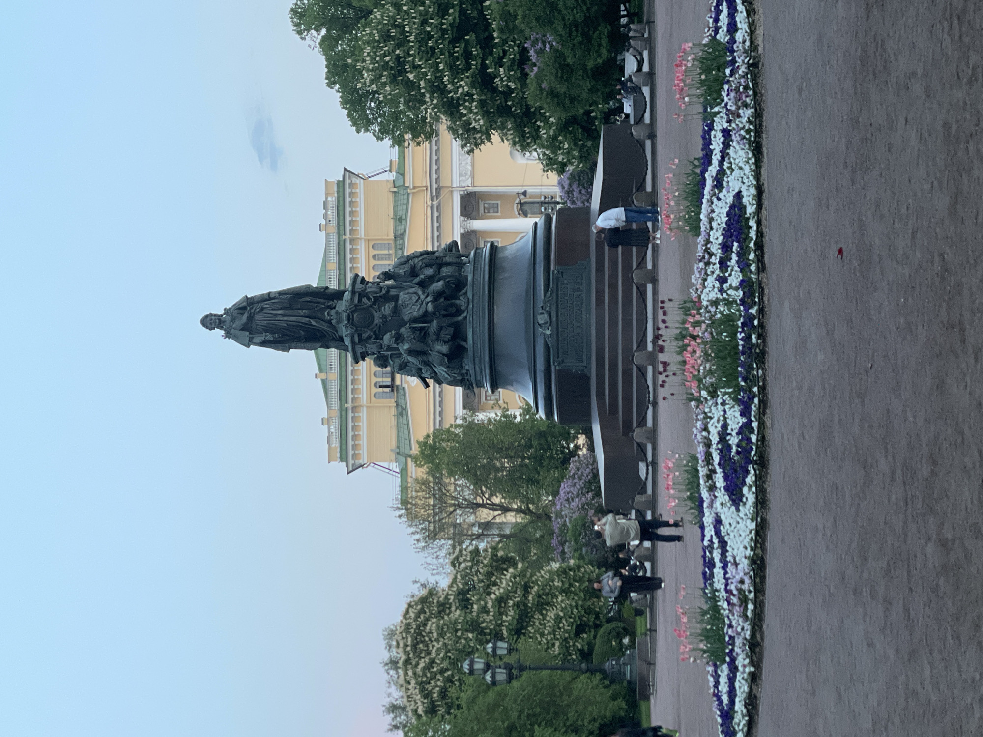 The image shows the Catherine the Great Monument, a bronze statue in Saint Petersburg, Russia. The statue is prominently displayed in a square, standing upon a large pedestal. It is surrounded by a flower bed with various colorful flowers. People are seen walking and standing around the monument, with some taking pictures. The background features a large building with a yellow facade and green roof. The sky is a pale blue, suggesting either early morning or late evening.