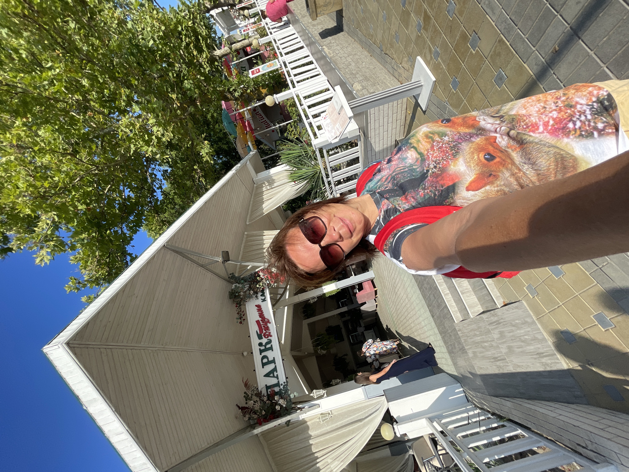 A woman is taking a selfie in front of a building with the sign "Парк Развлечений" (Park of Entertainment) in Anapa, Russia. The building appears to be a cafe or restaurant with white curtains and outdoor seating. The woman is wearing sunglasses and a t-shirt with a print of squirrels. The background shows trees, a blue sky, and a tiled walkway. There are other people visible in the background, including a woman in a blue dress and another in a pink dress. The scene is illuminated by bright sunlight.