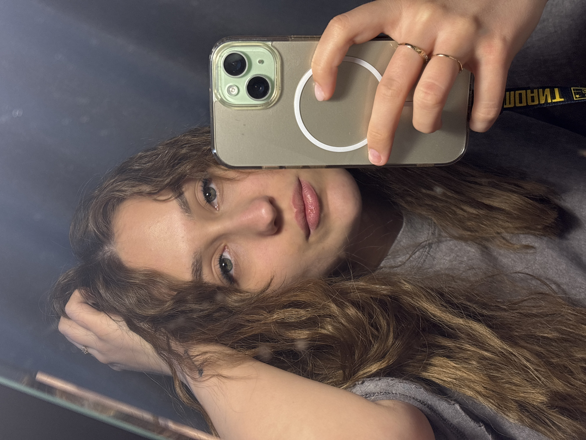 A young woman with wavy, light brown hair is taking a selfie using an iPhone with a light green case. The phone has a visible MagSafe ring. She is holding the phone up with her right hand and touching the screen with her index finger. She is wearing two rings on her right hand. Her left hand is near her head. She has fair skin, green eyes, and pink lips. She is wearing a grey shirt. A black and yellow lanyard or strap is visible near her right hand. The background appears to be a mirror reflecting the scene, with some blurry light reflecting off the surface. There is no evidence to suggest the image was taken in Марковский, Russia.