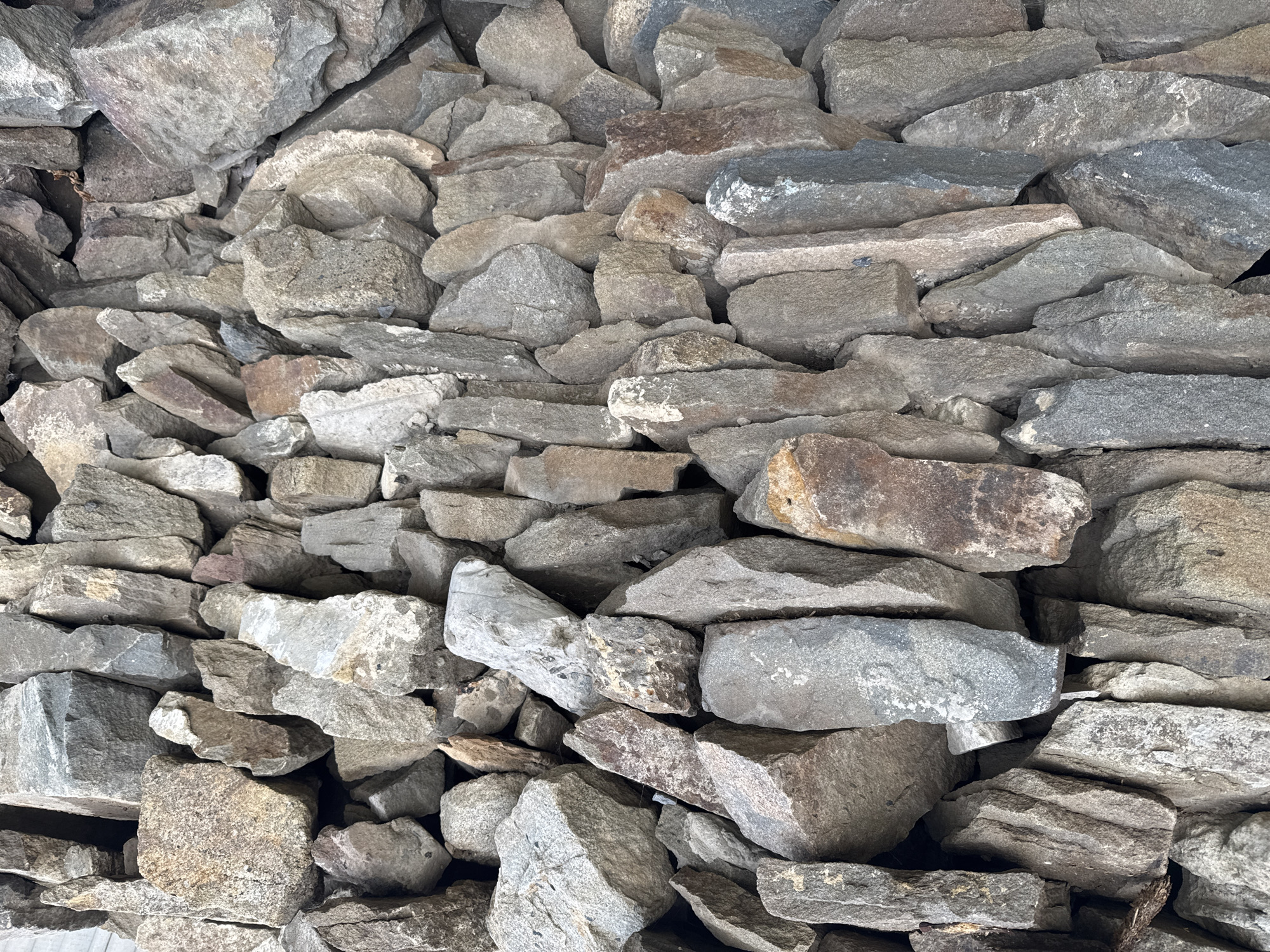 This media file displays a close-up, textured view of a rough-hewn stone wall. The stones are varied in size, shape, and color, ranging from light gray to darker grays and earthy browns, with some showing hints of reddish-brown. The surfaces of the stones are irregular, with many exhibiting natural cracks, rough textures, and chipped edges. They are stacked in an apparently haphazard but stable manner, creating a dense, solid wall. The lighting appears to be natural, possibly daylight, casting subtle shadows that accentuate the three-dimensional quality and depth of the stonework. There is no visible text, people, or any indication of a specific activity or event. The scene is purely a detailed study of the stone wall itself, suggesting it could be a detail of a building, boundary, or natural rock formation. The context provided places this image in Novoshakhtinsk, Russia.