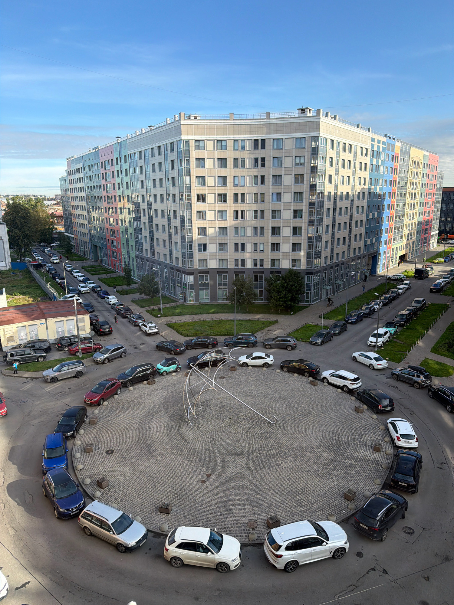 The image shows a view from above of a circular intersection with a modern residential building and parked cars. The building has a colorful facade with sections of pastel blue, green, pink, yellow, and red. The intersection is paved with cobblestones and features a contemporary sculpture in the center. Numerous cars are parked along the edge of the intersection and on the surrounding streets, forming lines of vehicles. The scene appears to be in a residential area of Saint Petersburg, Russia, likely a newly developed district, given the architecture and the orderly parking arrangement. There are a few pedestrians visible on the sidewalks and one person on a bicycle. The sky is blue with some clouds.