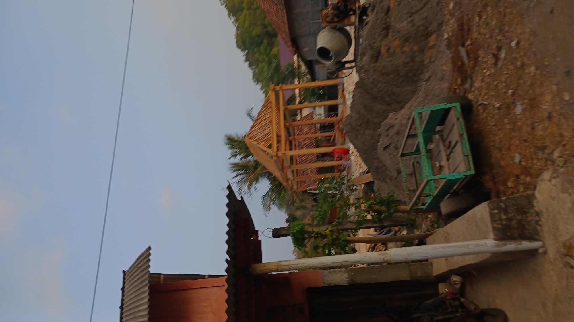 In the city of Tepus, Indonesia, an outdoor construction scene unfolds under a pale blue sky with sparse clouds. In the foreground, to the left, a portion of a building with a corrugated metal roof and an orange-colored wall is visible. A white concrete pillar supports the overhang. To the right of the building, a small green metal cart with two large wheels and a flatbed sits on reddish-brown dirt. Behind the cart, a large mound of dark gray construction material, likely sand or gravel, dominates the midground. Further back, a partially constructed wooden gazebo with a pitched roof structure is the central focus. The gazebo is supported by numerous wooden posts and beams, with some sections of a thatched roof visible. Several palm trees are present behind the gazebo, their fronds reaching towards the sky. To the far right, a cement mixer is positioned near a building with a reddish-brown tiled roof and dark gray walls. The ground throughout the scene is uneven and covered with dirt, small stones, and some scattered debris. The overall impression is one of ongoing construction or renovation in a semi-rural setting.