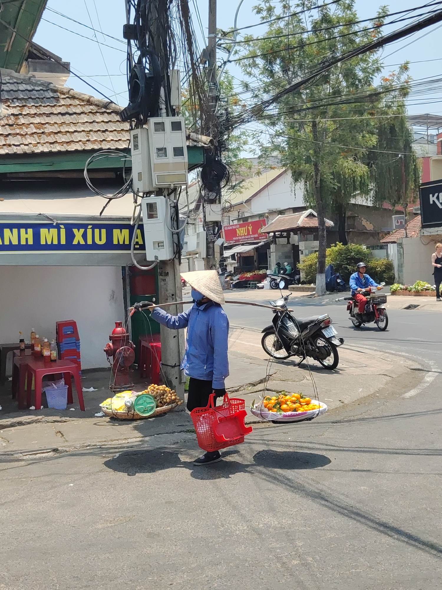 A street scene in Cam Ly - Đà Lạt, Vietnam, shows a woman wearing a conical hat, blue shirt, and black pants, balancing a shoulder pole with a red basket on one side and a basket of yellow fruit and a small scale on the other. She stands on the sidewalk, next to a building with a sign in yellow Vietnamese script, and a red fire hydrant. The street is visible in the background with motorbikes and a person on a moped. Power lines are densely tangled above. Another building is visible behind, with a sign in red Vietnamese script. A person is visible on the far right.