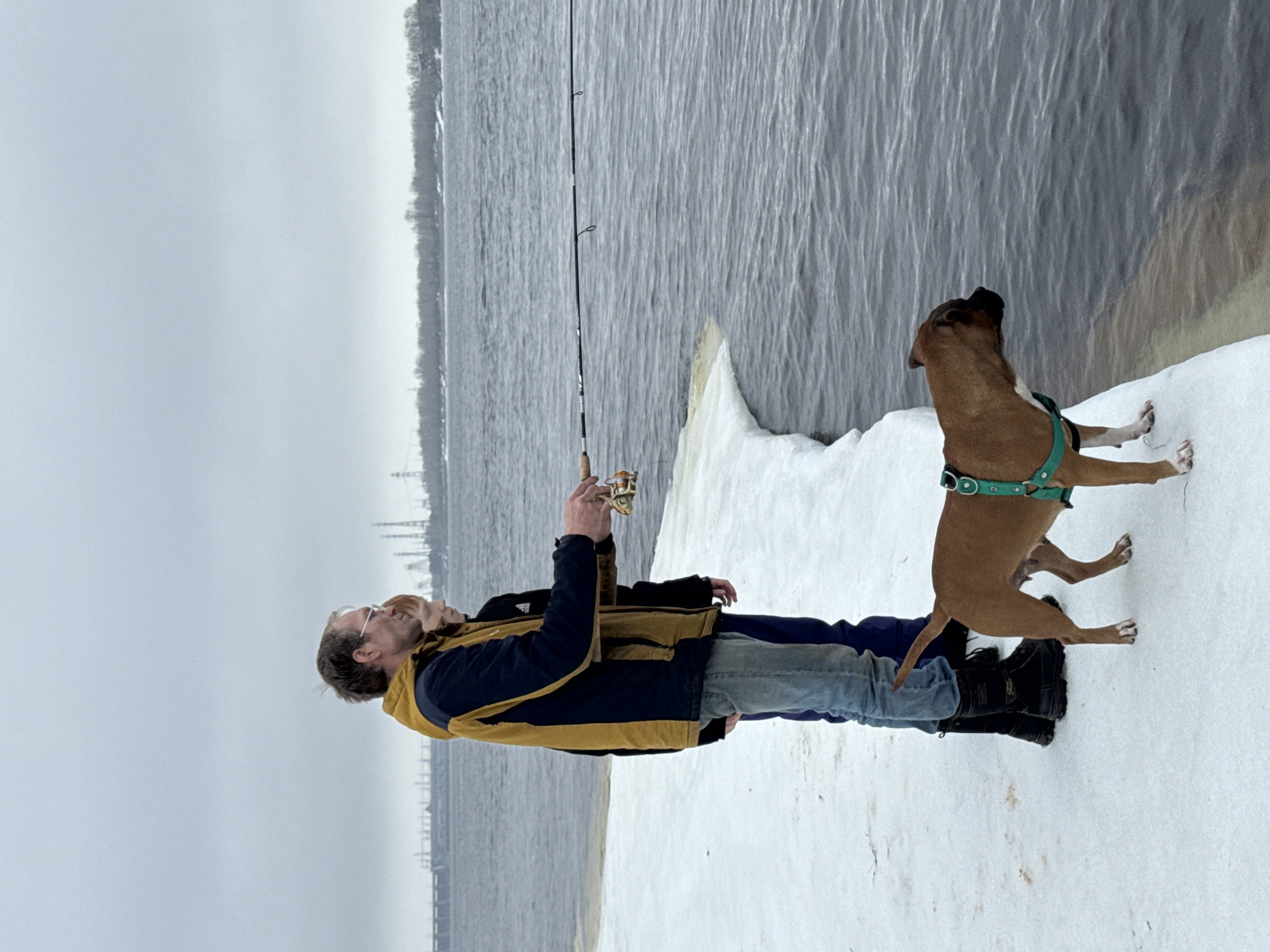 A man and a dog stand on a snowy bank next to a body of water in Chaikovsky, Russia. The man holds a fishing rod. The dog, wearing a green harness, stands on its hind legs looking up and toward the water. The water is calm with a few ripples. A hazy sky is overhead. The shoreline of Chaikovsky can be seen across the water.