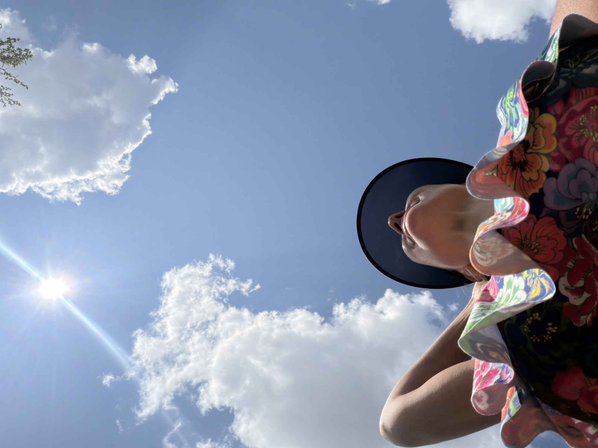 A low-angle shot shows a person looking up at a bright sun in a clear blue sky with scattered white clouds. The person, wearing a wide-brimmed hat and a colorful floral top, has one arm behind their head. The image was taken on a sunny day, possibly in the city of Novosibirsk, Russia, given the angle of the sun suggesting it is midday or early afternoon in a northerly latitude.