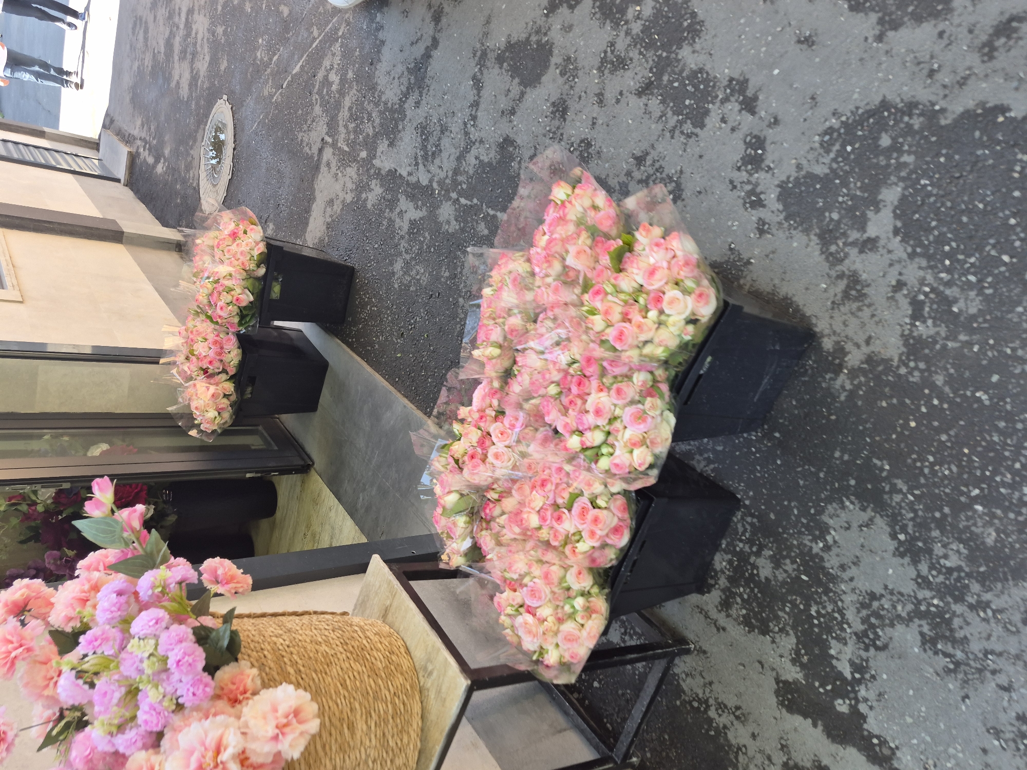 This image captures a vibrant display of pink flowers outside a flower shop, possibly in Yerevan, Armenia. In the foreground, several large bouquets of pink roses are arranged in black plastic crates on the asphalt ground. The roses are tightly packed and appear fresh, with cellophane wrapping protecting some of them. To the left, a woven basket overflowing with larger pink carnation-like flowers sits on a wooden table with a metal frame.
In the background, the entrance to the flower shop is visible. Through the glass door, more flowers, including darker red and purple blooms, can be seen. Black planters are also present inside the shop. The exterior of the building is a light-colored stucco.
The ground is paved with a dark asphalt surface that shows signs of wear and is wet in patches, reflecting the sunlight. A circular metal grate is embedded in the asphalt near the flower displays. In the far background, two individuals are walking away from the shop, their lower legs and feet visible, suggesting a sunny day. The overall impression is that of a bustling flower shop with an attractive outdoor display.
