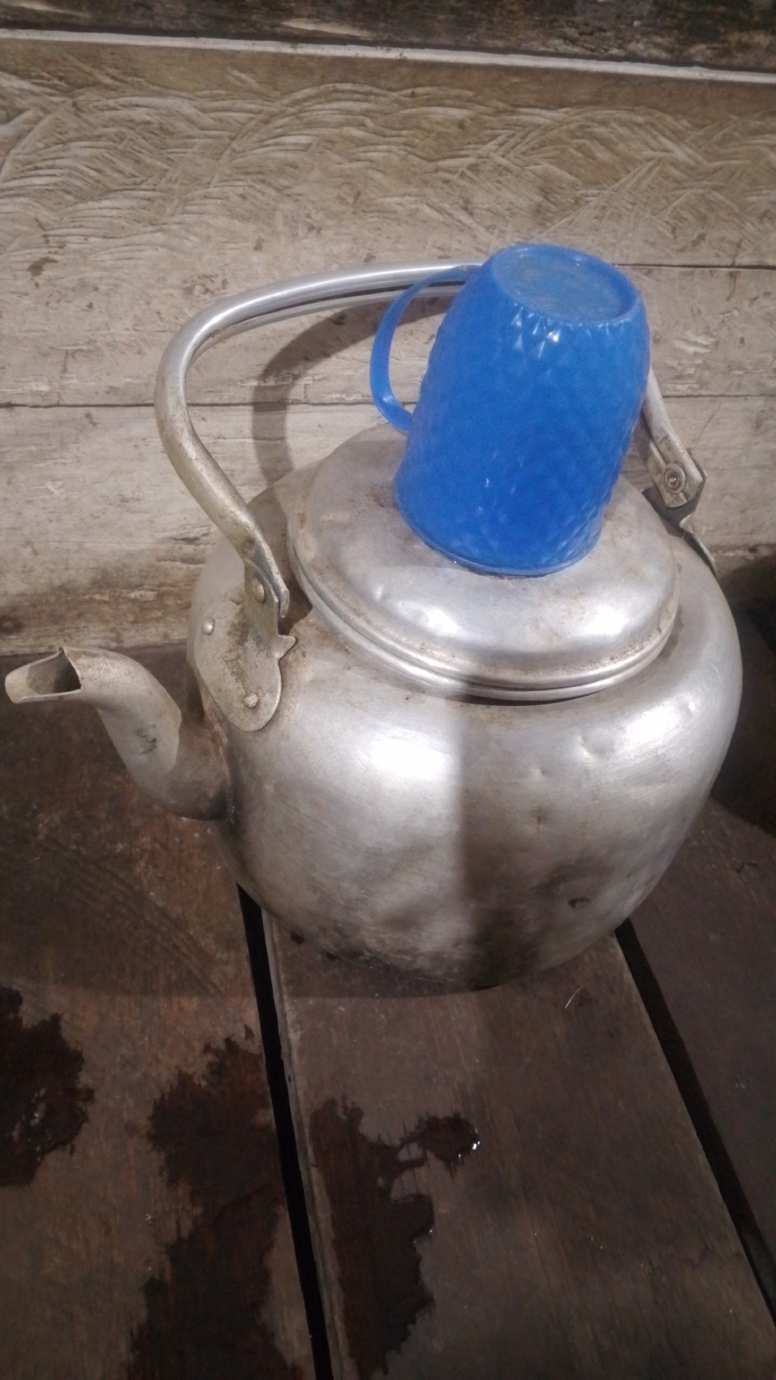 A metallic kettle with a silver handle and spout rests on a wooden surface. A blue plastic cup is inverted and placed on top of the kettle's lid. The wooden surface appears worn and has some dark stains, possibly from water or other liquids. The background consists of a wooden wall with visible planks and grain. The lighting suggests an indoor or covered outdoor setting, possibly in a kitchen or storage area. The image does not contain any elements that would allow for a specific location in Olak-Olak Kubu, Indonesia to be identified.