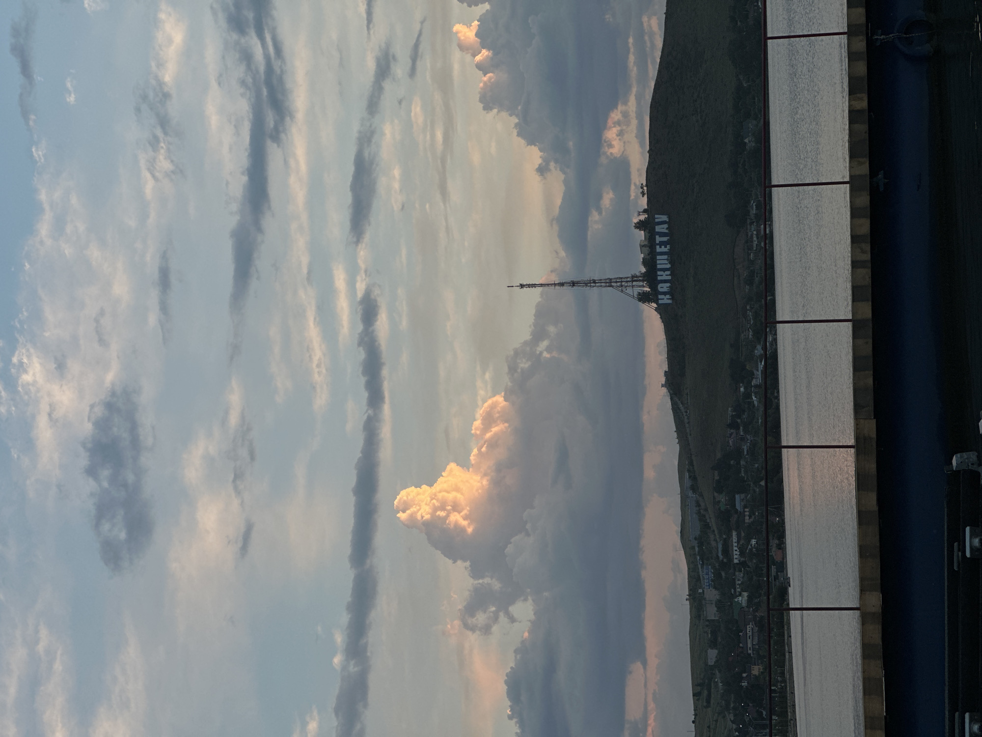 The image depicts a scenic view of the city of Kokshetau, Kazakhstan, likely taken from across a body of water. In the foreground, a dark blue structure, possibly a pier or embankment, with a railing made of red metal poles and horizontal bars, spans the bottom of the frame. Beyond this barrier, a calm body of water reflects the muted colors of the sky.
The middle ground is dominated by a large, grassy hill. At the summit of the hill, a tall, slender radio tower stands prominently. Below and to the right of the tower, large white letters spelling out "КОКШЕТАУ" (Kokshetau) are visible against the dark hillside. A scattering of trees and buildings can be seen on the slopes of the hill, suggesting a town or settlement. The background is filled with a dramatic sky, characterized by a mix of dark, heavy clouds and lighter, illuminated clouds catching the warm light of either sunrise or sunset. One particularly large, illuminated cloud formation stands out on the left side of the frame. The overall impression is one of a tranquil evening or morning scene in Kokshetau.