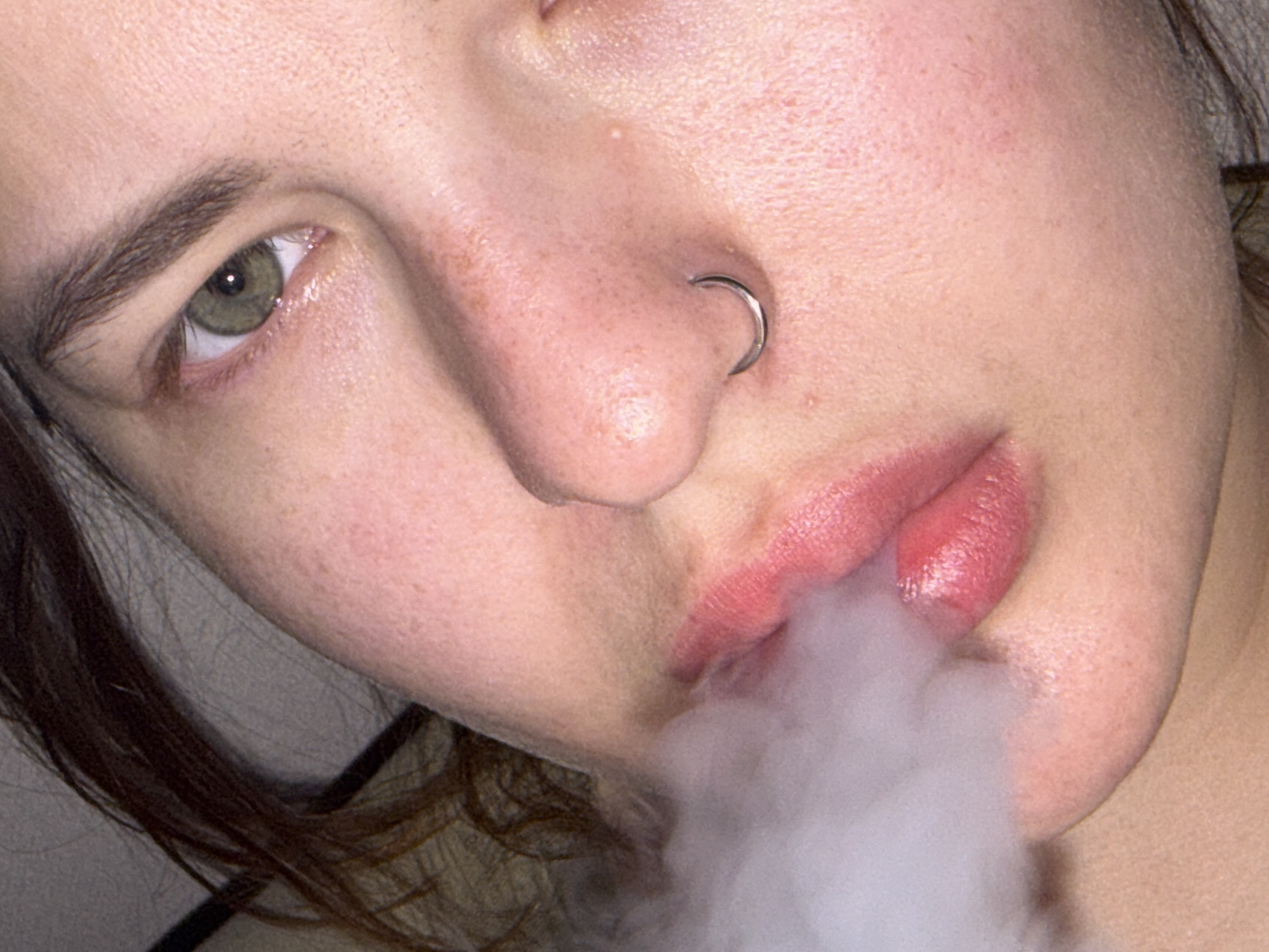 A close-up image shows a person's face angled toward the camera. The person has fair skin, a nose ring, and pink lips from which a cloud of white smoke or vapor is emanating. The person has one eye visible with green irises, and dark eyebrows. The environment appears to be undefined.