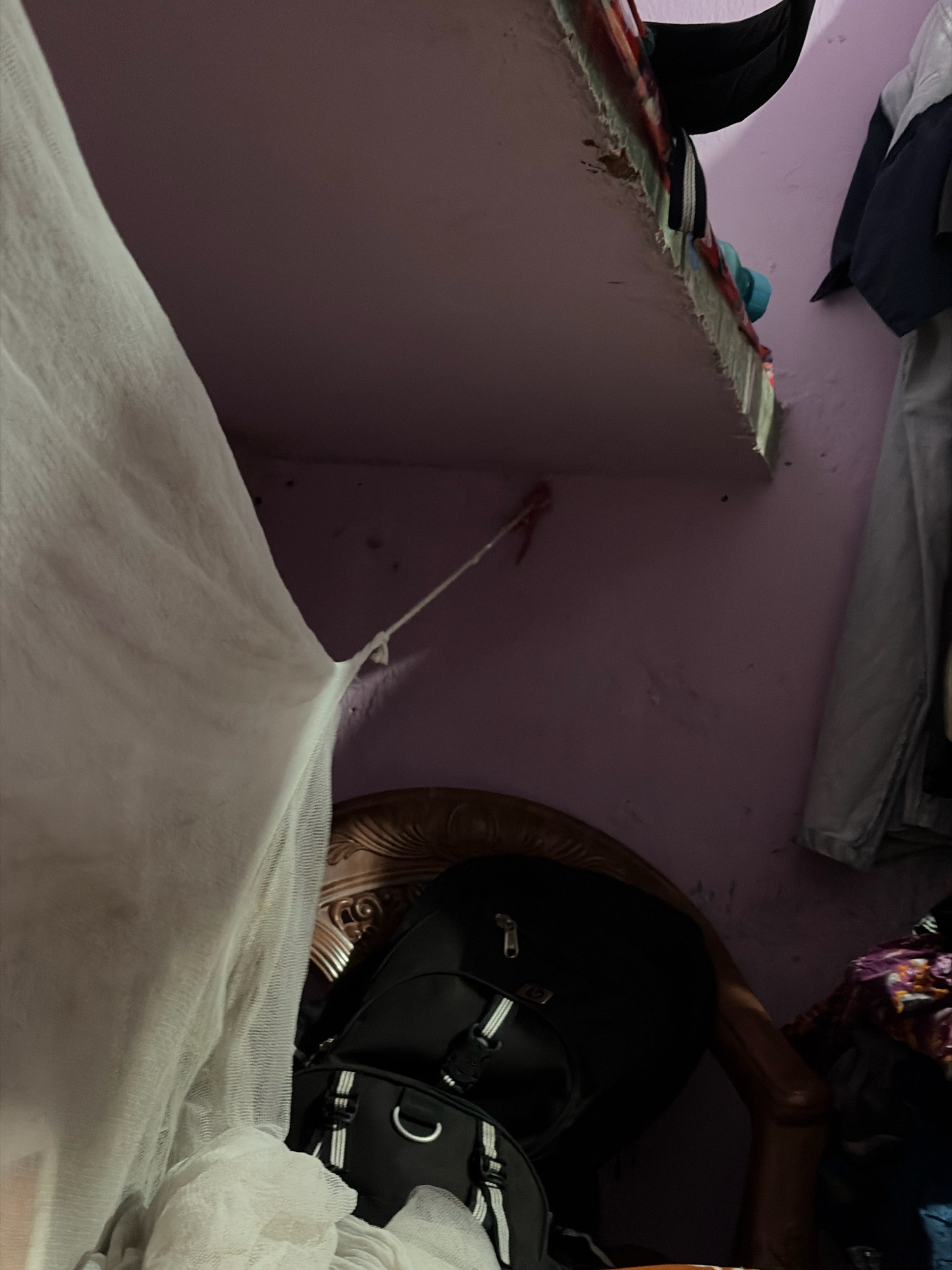 The image depicts a cluttered corner of a room. A white sheer fabric, possibly a mosquito net, hangs prominently on the left side, extending towards the upper left quadrant and obscuring parts of the scene. A string is attached to this fabric, tied to the pink-colored wall in the center.
Above, a shelf is visible with various items, including clothing with colorful patterns, a black object, and a teal-colored item. The shelf appears to be slightly damaged at the edge. To the right of the shelf, clothing hangs on the wall, including a navy blue jacket and a light gray garment.
In the lower center of the image, a black backpack is positioned on what appears to be a wooden chair with a decorative, ornate backrest. The backpack has white striped accents and metal hardware. There are also folded white fabrics or textiles below the backpack.
The overall environment suggests a residential space, possibly a bedroom or storage area, with items piled and hanging. The lighting is dim, casting shadows and contributing to the cluttered aesthetic. The location is identified as Jamshedpur, India.