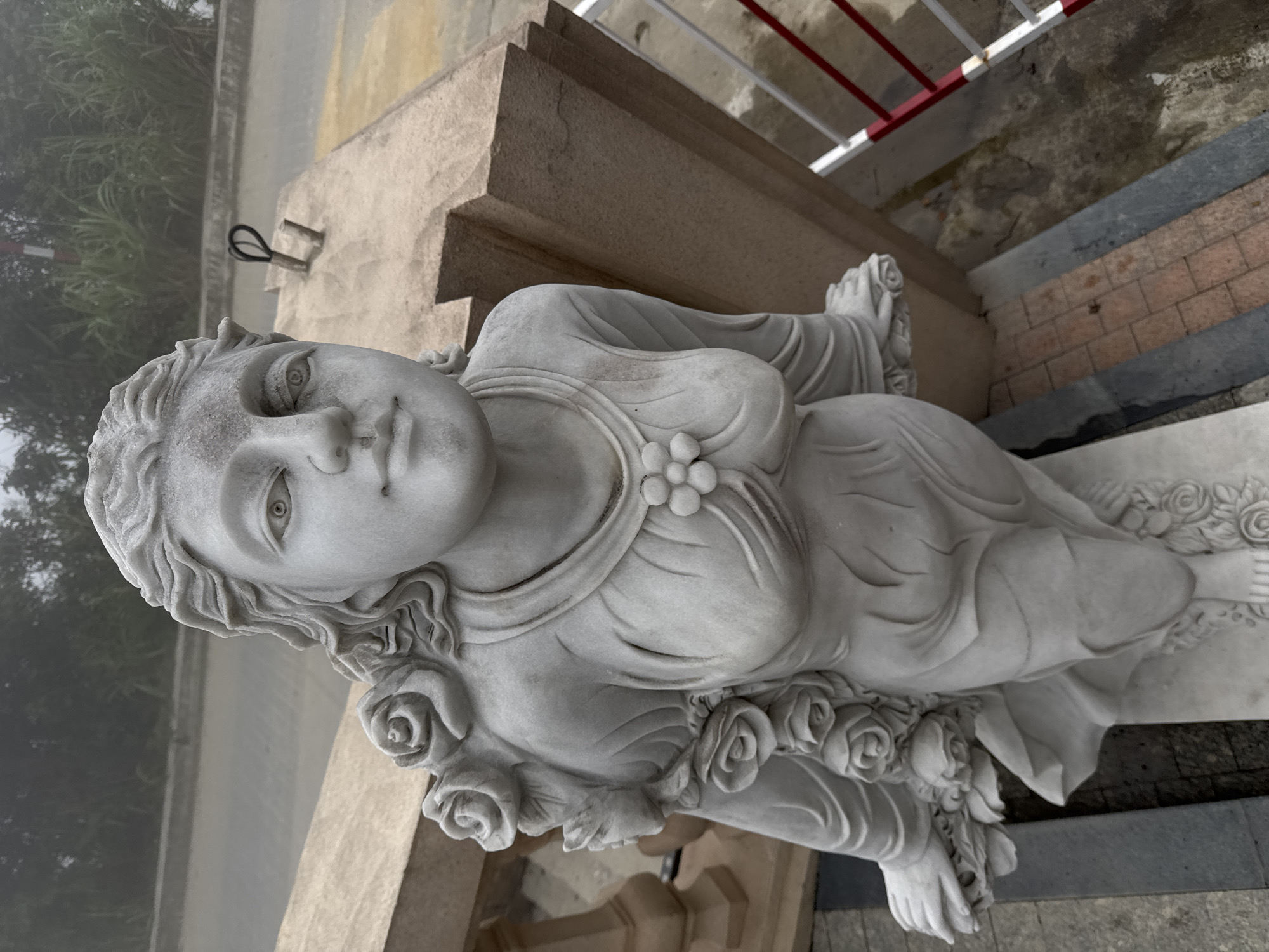 A marble statue of a woman, adorned with floral garlands, is positioned near a set of concrete steps with a white and red railing. The statue appears weathered and is placed outdoors. In the background, a foggy or misty scene blurs the surroundings, suggesting a location in a high-altitude area. The presence of a road or path in the background suggests the statue is in a public area or garden. The location is possibly in the Bà Nà Hills, Đà Nẵng, Vietnam.