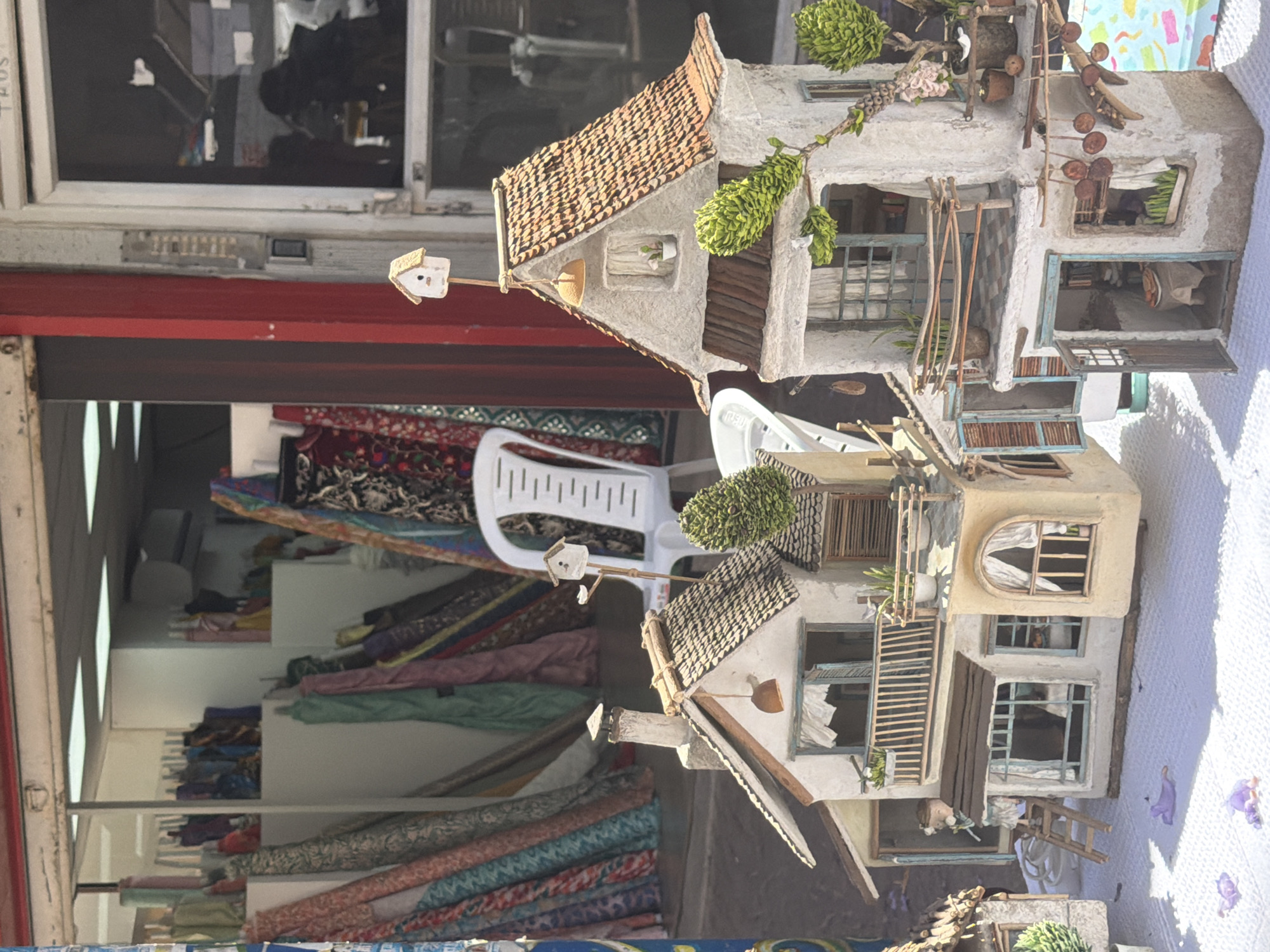 A horizontally oriented image shows a small, intricate model of a house displayed outside a shop. The shop, on the left, is filled with rolls of fabric of various colors and patterns stacked on shelves. A white plastic chair sits amidst the fabric. The model house is positioned to the right of the shop entrance, partially resting on a white surface. The model is highly detailed, featuring a multi-tiered structure with faux stone walls, tiled roofs, miniature windows, balconies, and decorative elements like small trees and wooden ladders. A tiny birdhouse is mounted on a pole extending from one of the roofs. The background suggests an urban setting. The location is Tel-Aviv, Israel.