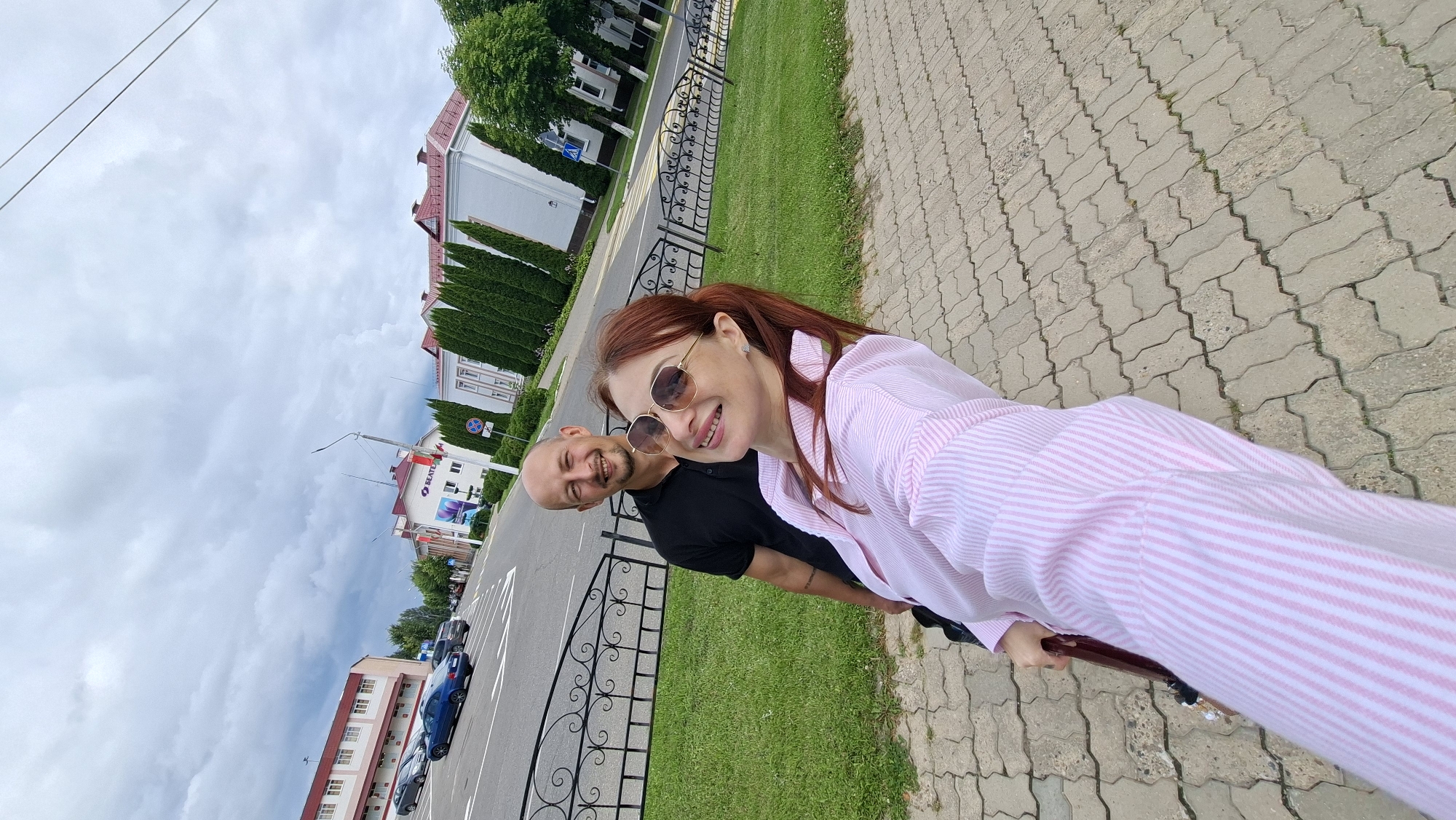In the city of Myadzyel, Belarus, a man and a woman are photographed outdoors, likely on a street or sidewalk, with buildings and a road visible in the background. The woman, in the foreground, is wearing a pink and white striped shirt and sunglasses, and appears to be taking a selfie. She is smiling and looking towards the camera. Behind her, the man, wearing a black polo shirt, is also smiling and looking at the camera. He has a shaved head. A black metal fence separates the sidewalk from a grassy area and the road. The buildings in the background appear to be commercial or institutional, with a sign that reads "BEATE." The sky is overcast, with visible clouds. The overall scene suggests a casual outing or visit to the town.