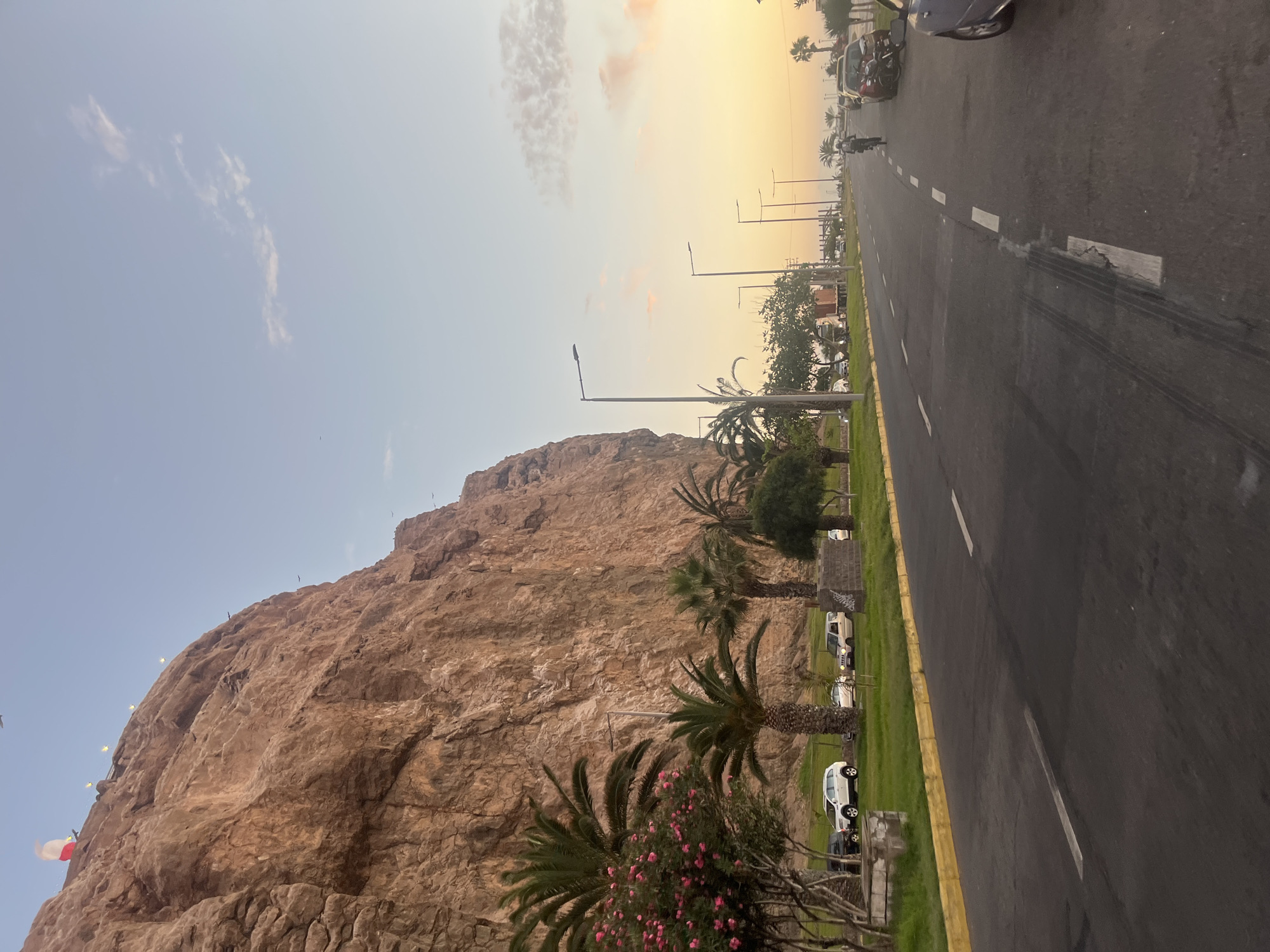 The image depicts a view of El Morro de Arica, a prominent cliff overlooking the city of Arica, Chile. The foreground shows a road running alongside the base of the cliff, lined with palm trees and some flowering bushes. Several vehicles are parked on the roadside. The road stretches into the distance, where a few cars and a motorcycle are visible. Overhead, the sky transitions from a light blue to a gradient of yellow and orange towards the horizon, indicating either sunrise or sunset. A few clouds are scattered across the sky.
