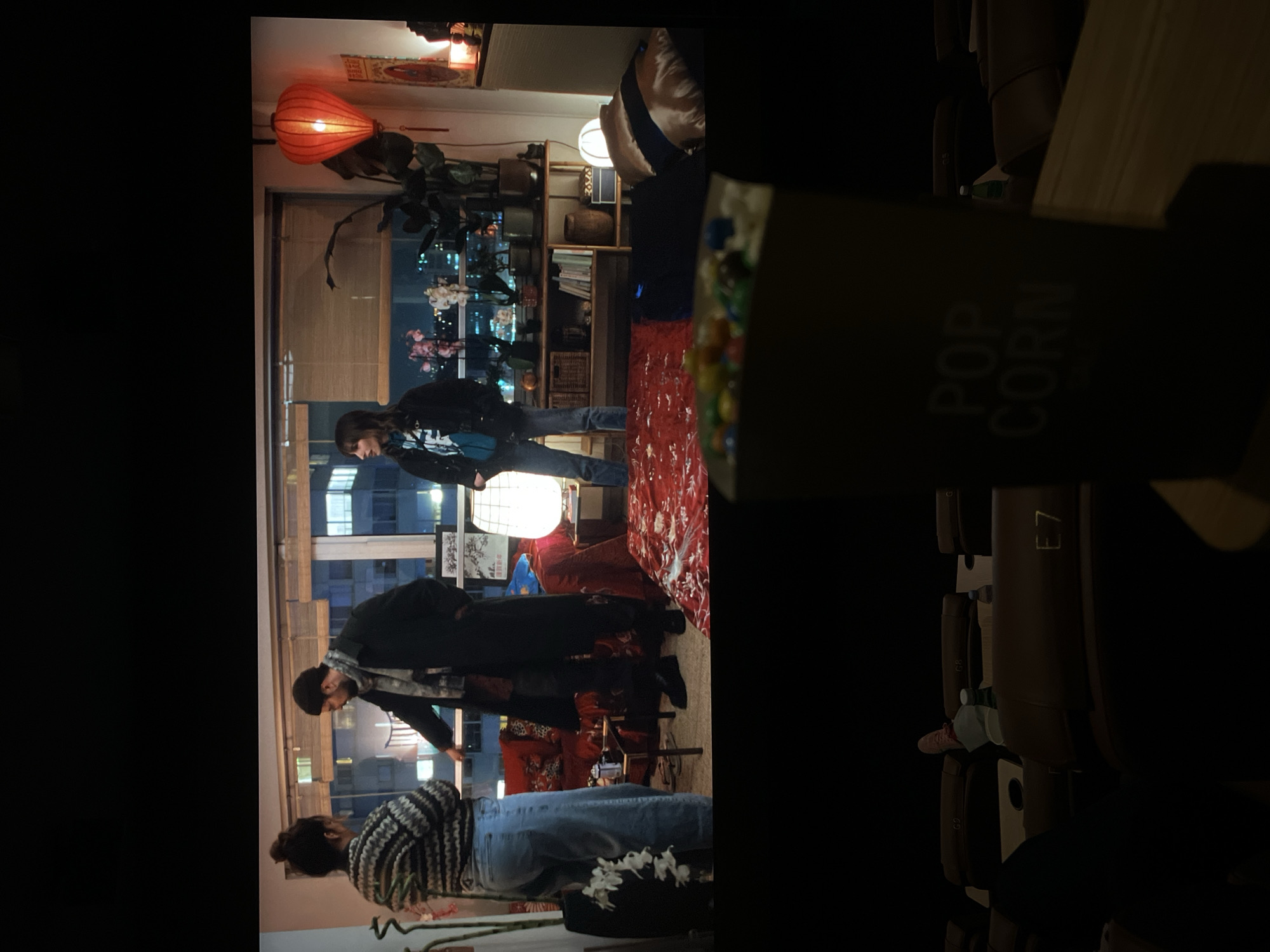 The image shows the interior of a cinema or home theater, with rows of leather chairs visible in the background. A container with the word "POPCORN" on it is prominently featured in the foreground, partially obscuring the view of the room.
Through a large window in the background, a cityscape is visible, suggesting the location could be Paris. Several people are inside the room. One is wearing a striped sweater and jeans, while another is in a black jacket. They seem to be standing near a red patterned rug and a lamp. The overall lighting is dim. There are shelves and plants near the window.