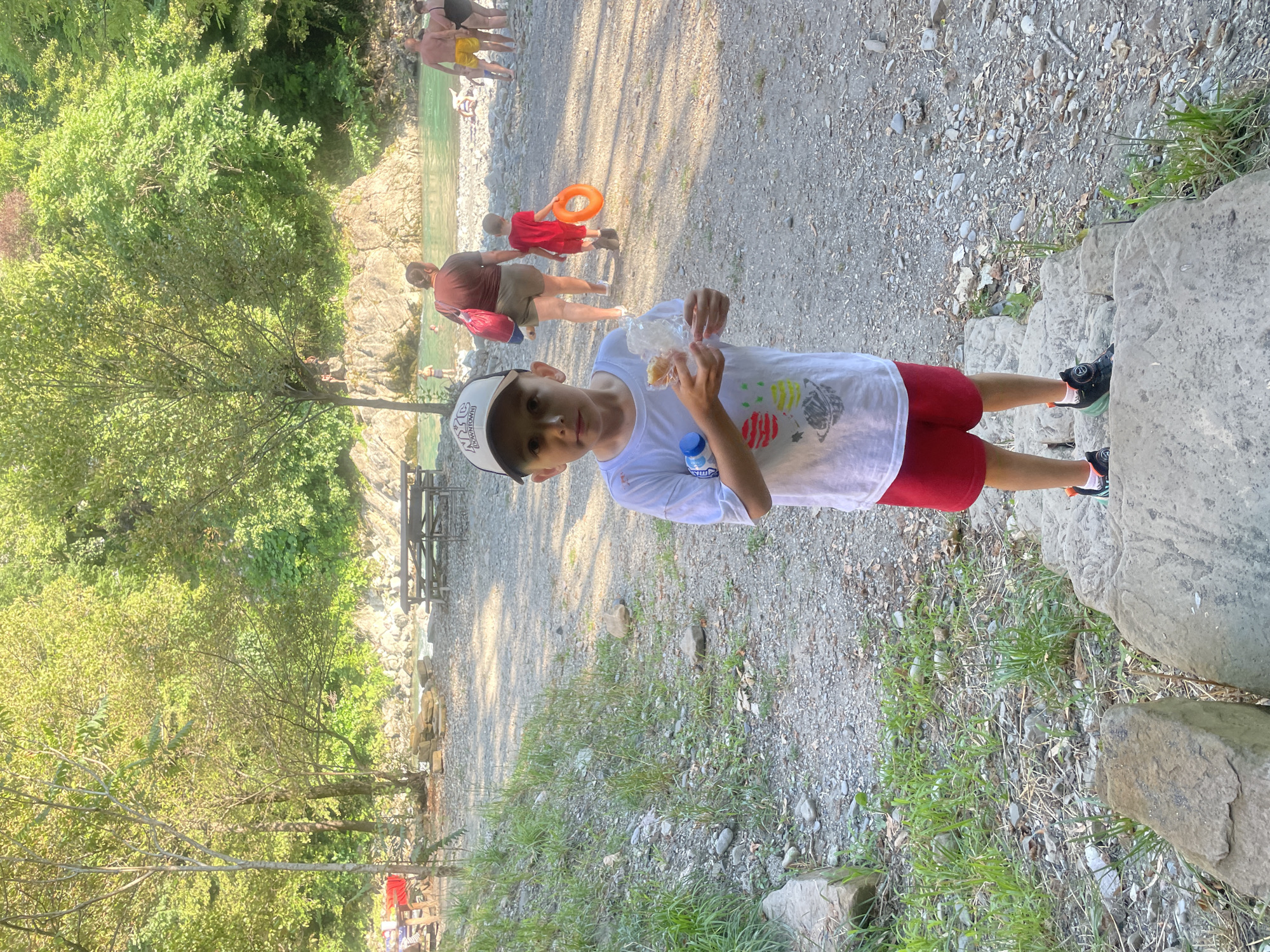 In Sochi, Russia, a young boy stands on a stone pathway leading down to a riverbank. He wears a white t-shirt with planet graphics, red shorts, a baseball cap, and sneakers. He holds a snack in a plastic bag and a bottle of water. In the background, people are visible near the turquoise water of a river. Some are wading, while others are relaxing on the shore. Trees and rocks line the riverbank, creating a natural, outdoor recreational area. Picnic tables and benches are scattered in the vicinity, suggesting a place for gatherings. The sun is shining, casting a warm light on the scene.